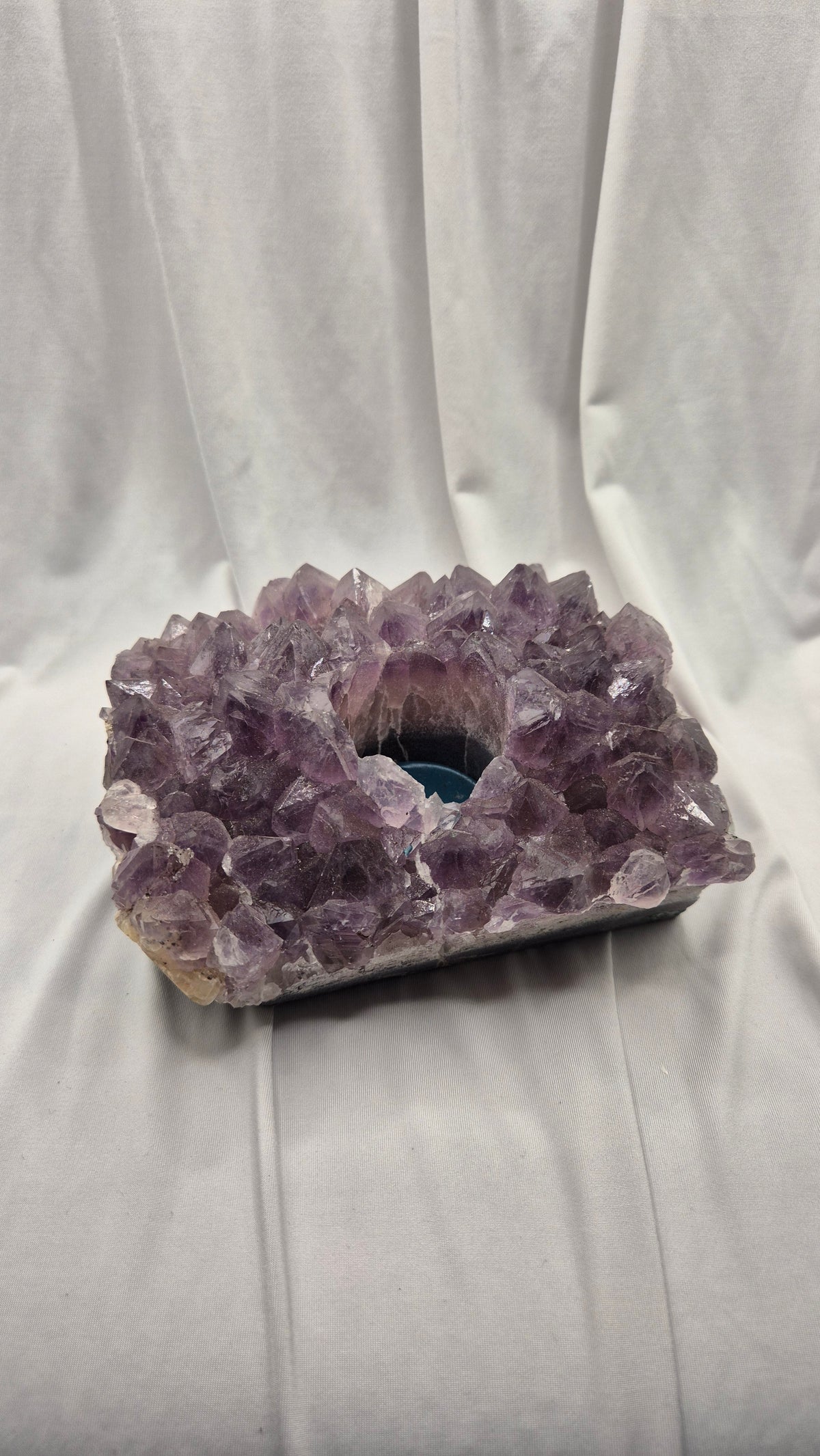 A raw amethyst crystal cluster geode with a hollow center, resting on a white draped fabric background.
