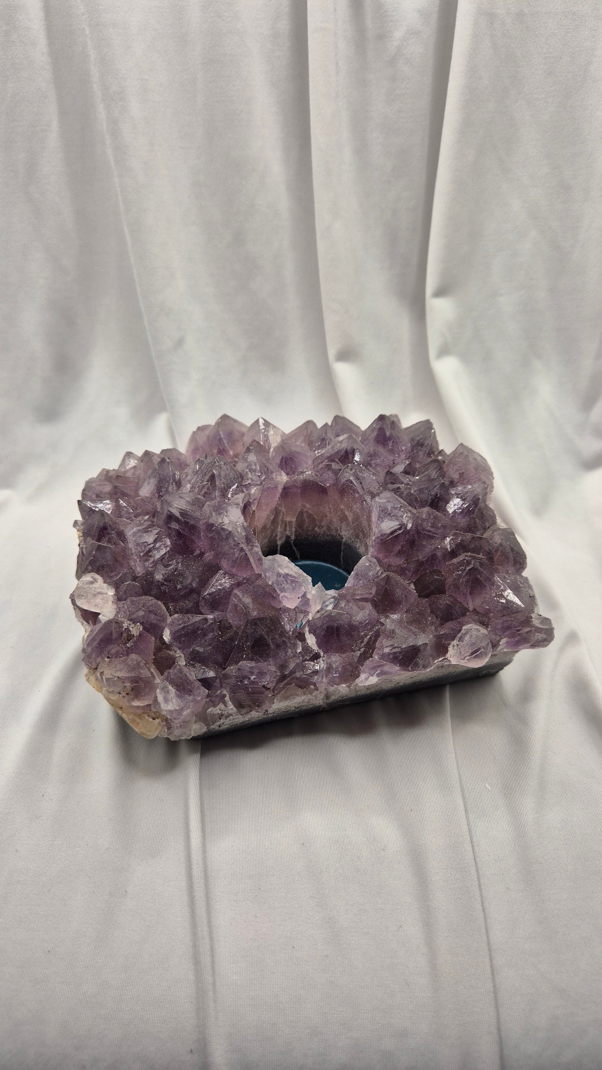 A raw amethyst crystal cluster geode with a hollow center, resting on a white draped fabric background.