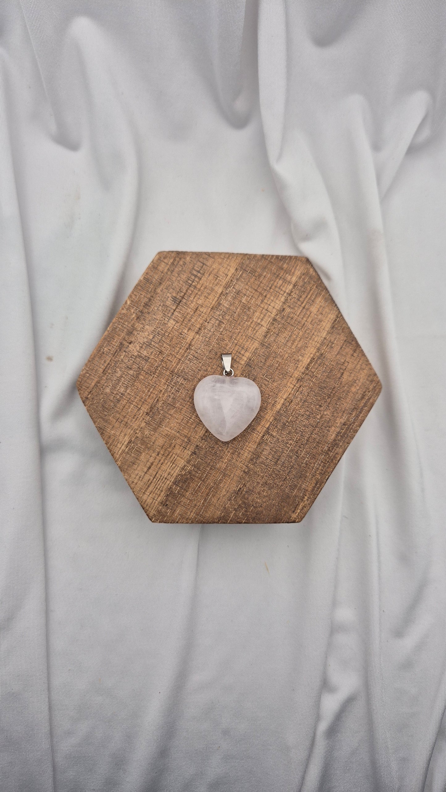 A heart-shaped rose quartz pendant on a silver bail, displayed on a hexagonal wooden block over a draped white fabric background.