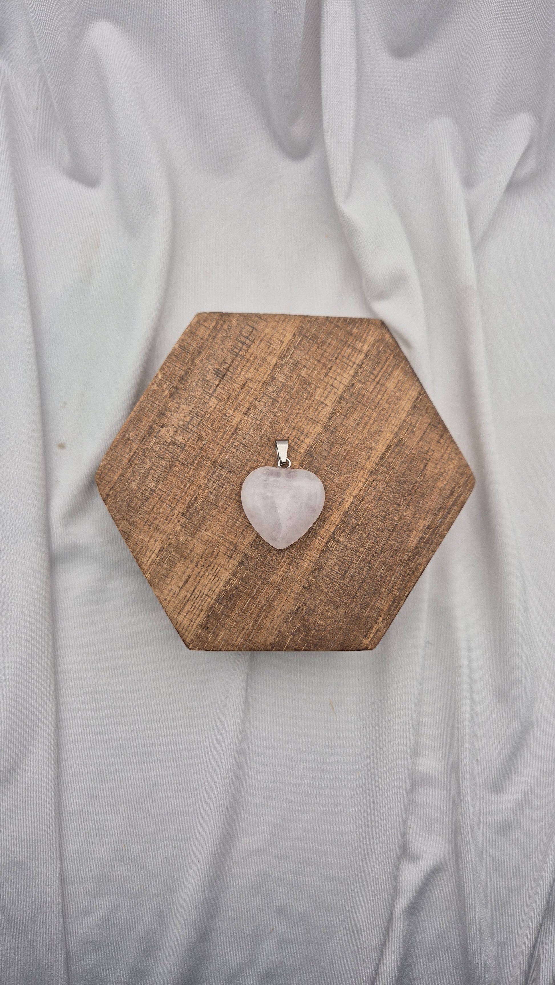 A heart-shaped rose quartz pendant on a silver bail, displayed on a hexagonal wooden block over a draped white fabric background.