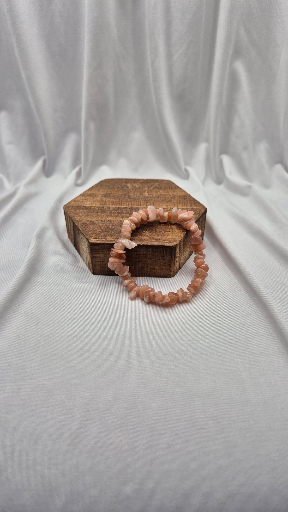 A peach-colored beaded stretch bracelet made of rough, irregular stone pieces, displayed on a small hexagonal wooden box against a draped white fabric background.
