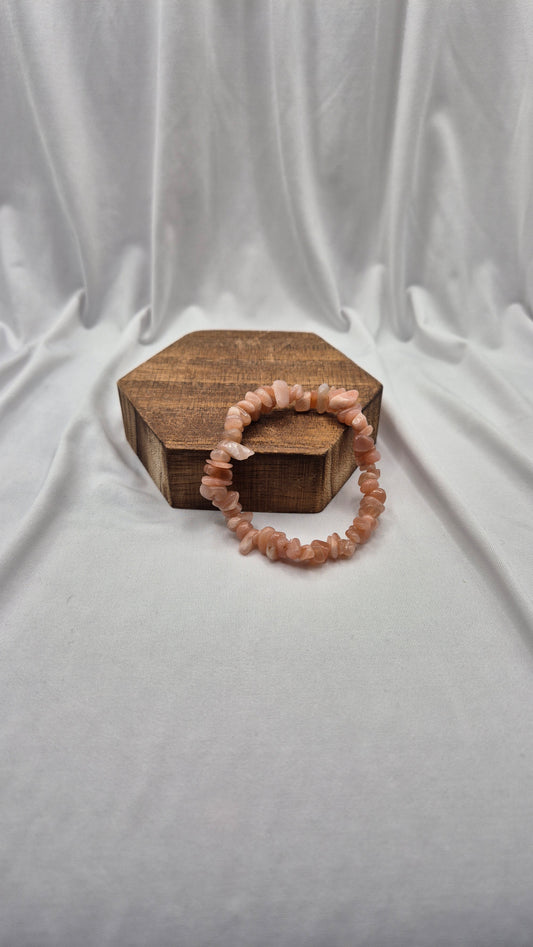 A peach-colored beaded stretch bracelet made of rough, irregular stone pieces, displayed on a small hexagonal wooden box against a draped white fabric background.