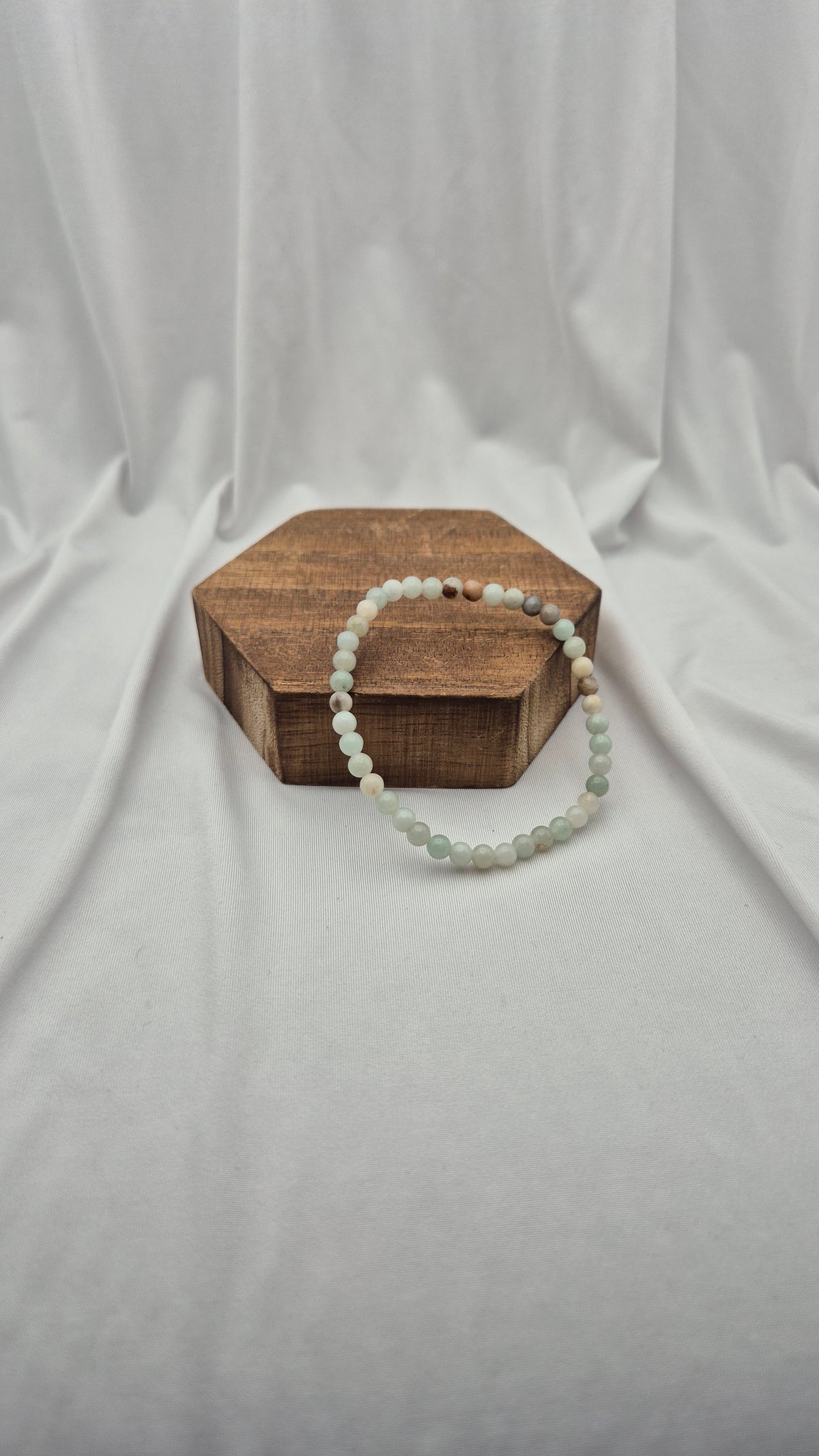 A beaded bracelet made of pale green and white round beads, draped over a hexagonal wooden block against a draped white fabric backdrop.