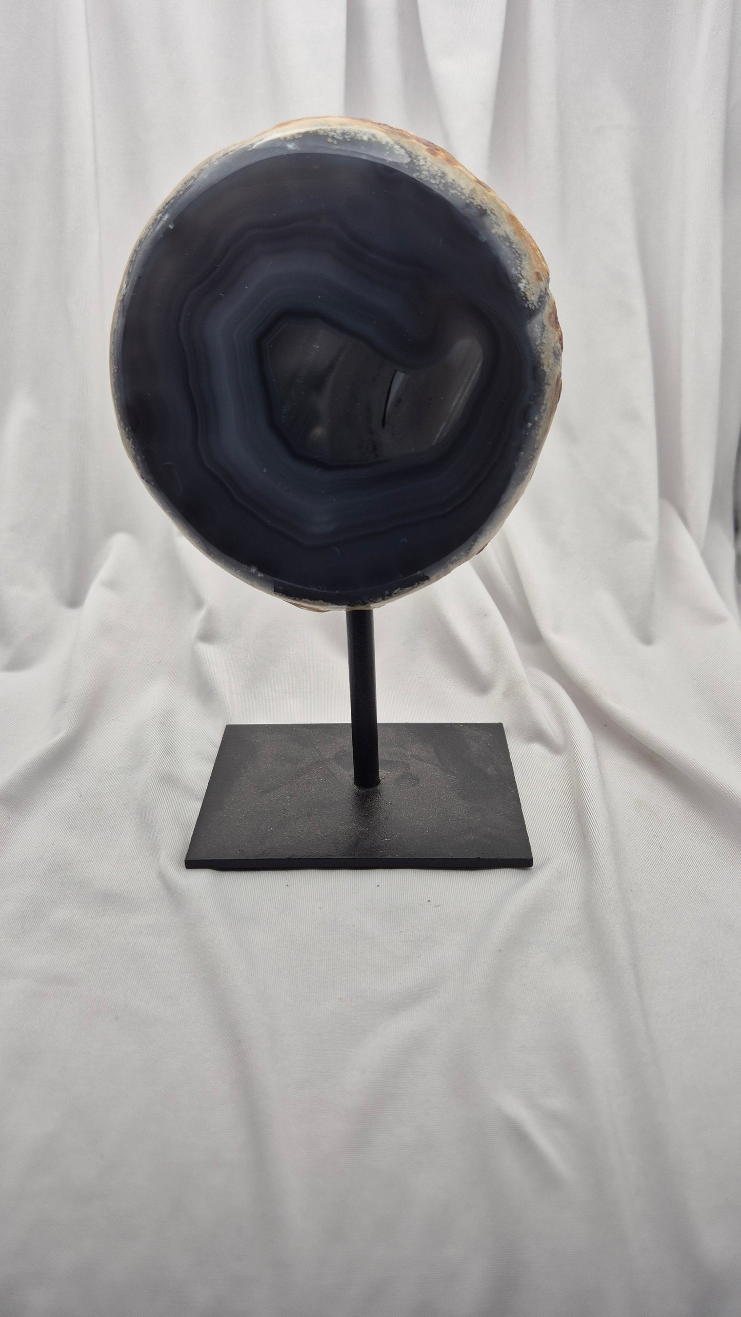 A polished agate geode slice with dark blue, black, and gray banded patterns and a light tan outer rim, displayed on a black metal stand against a draped white fabric background.