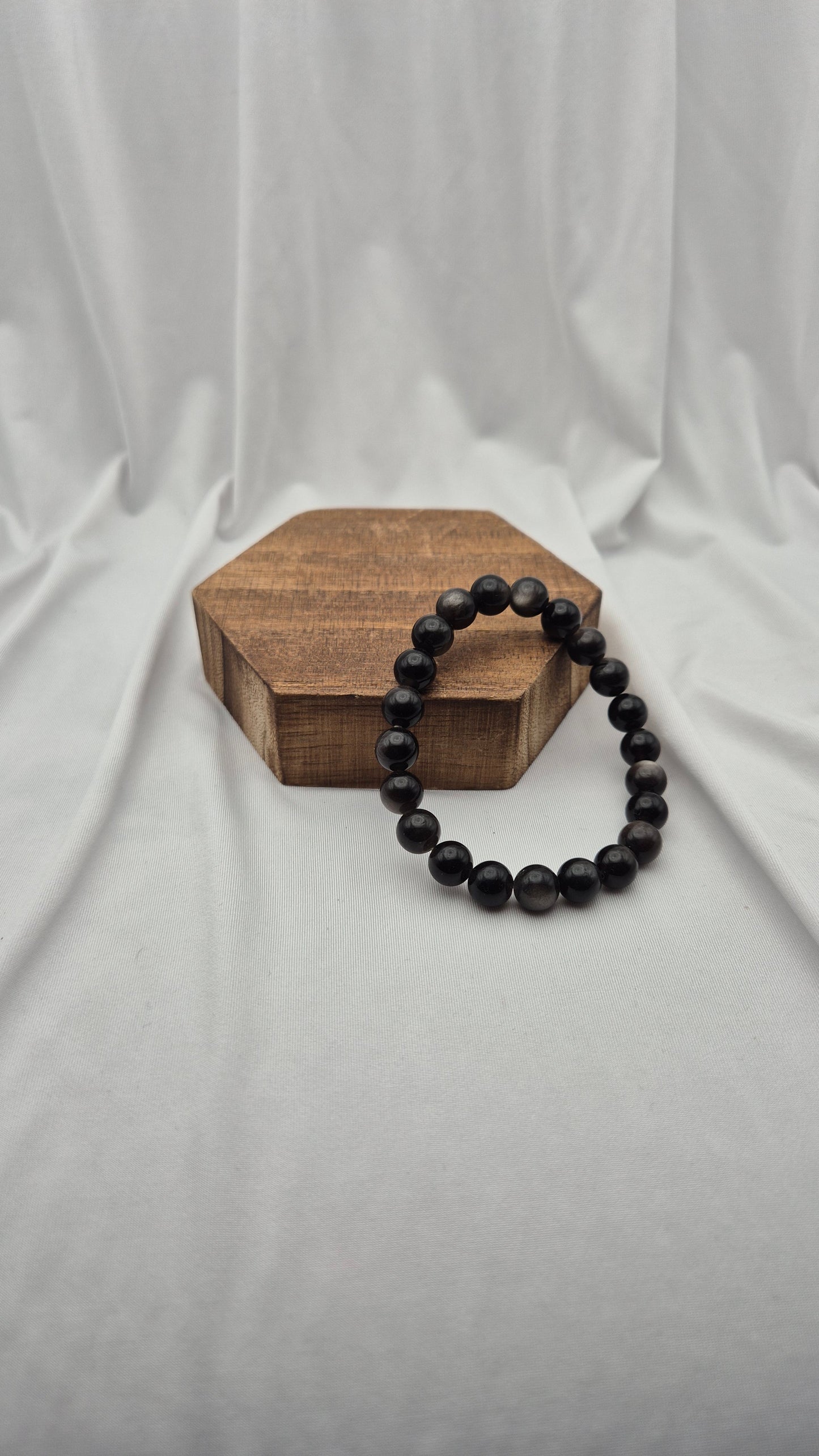 A black beaded bracelet draped over a hexagonal wooden block, set against a draped white fabric backdrop.