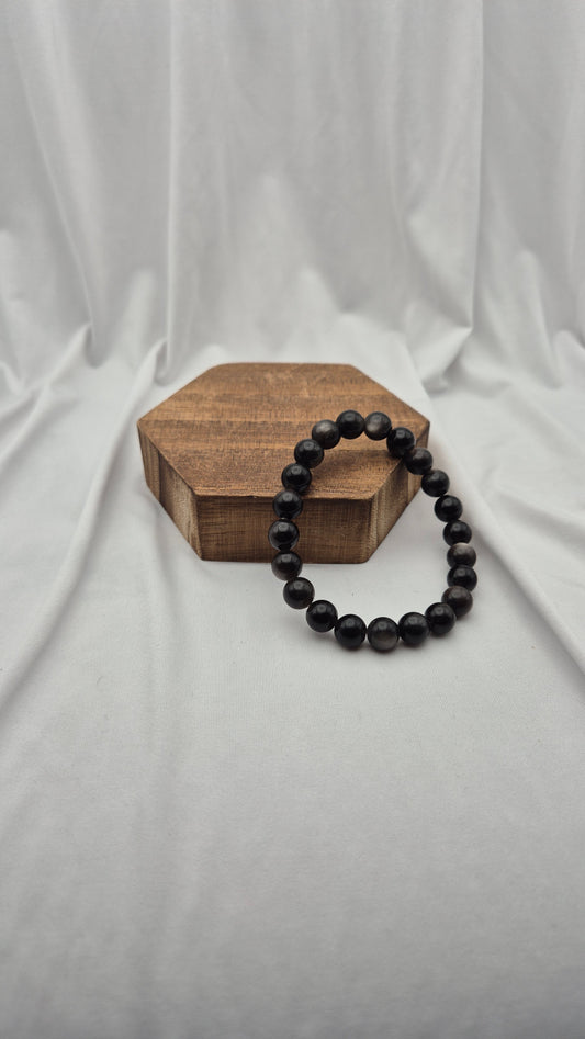 A black beaded bracelet draped over a hexagonal wooden block, set against a draped white fabric backdrop.