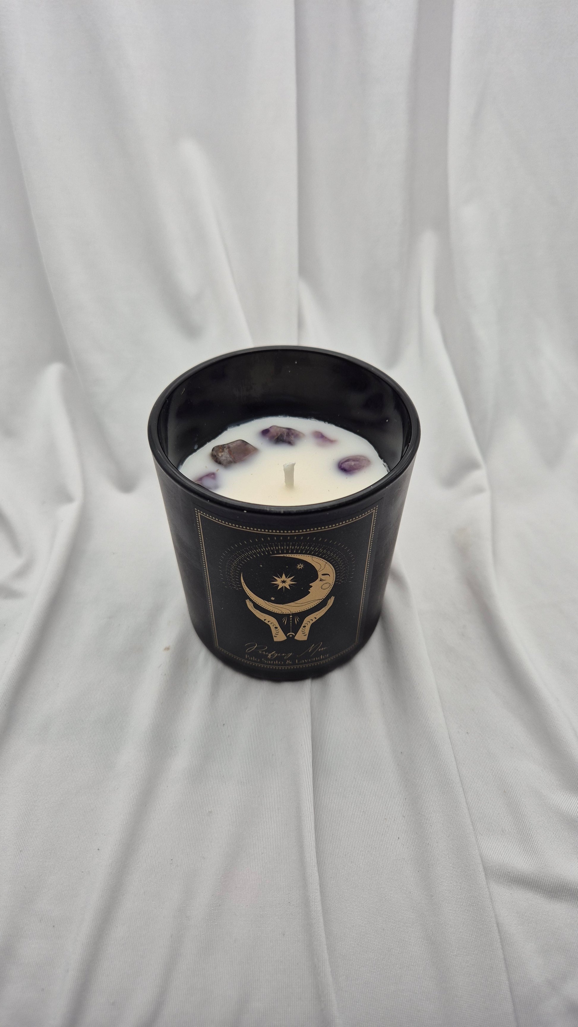 A black glass jar candle with white wax and floating dried botanical pieces, featuring a gold celestial graphic, set against a draped white fabric background.