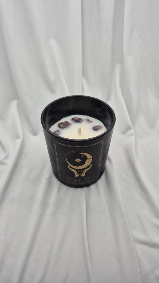 A black glass jar candle with white wax and floating dried botanical pieces, featuring a gold celestial graphic, set against a draped white fabric background.