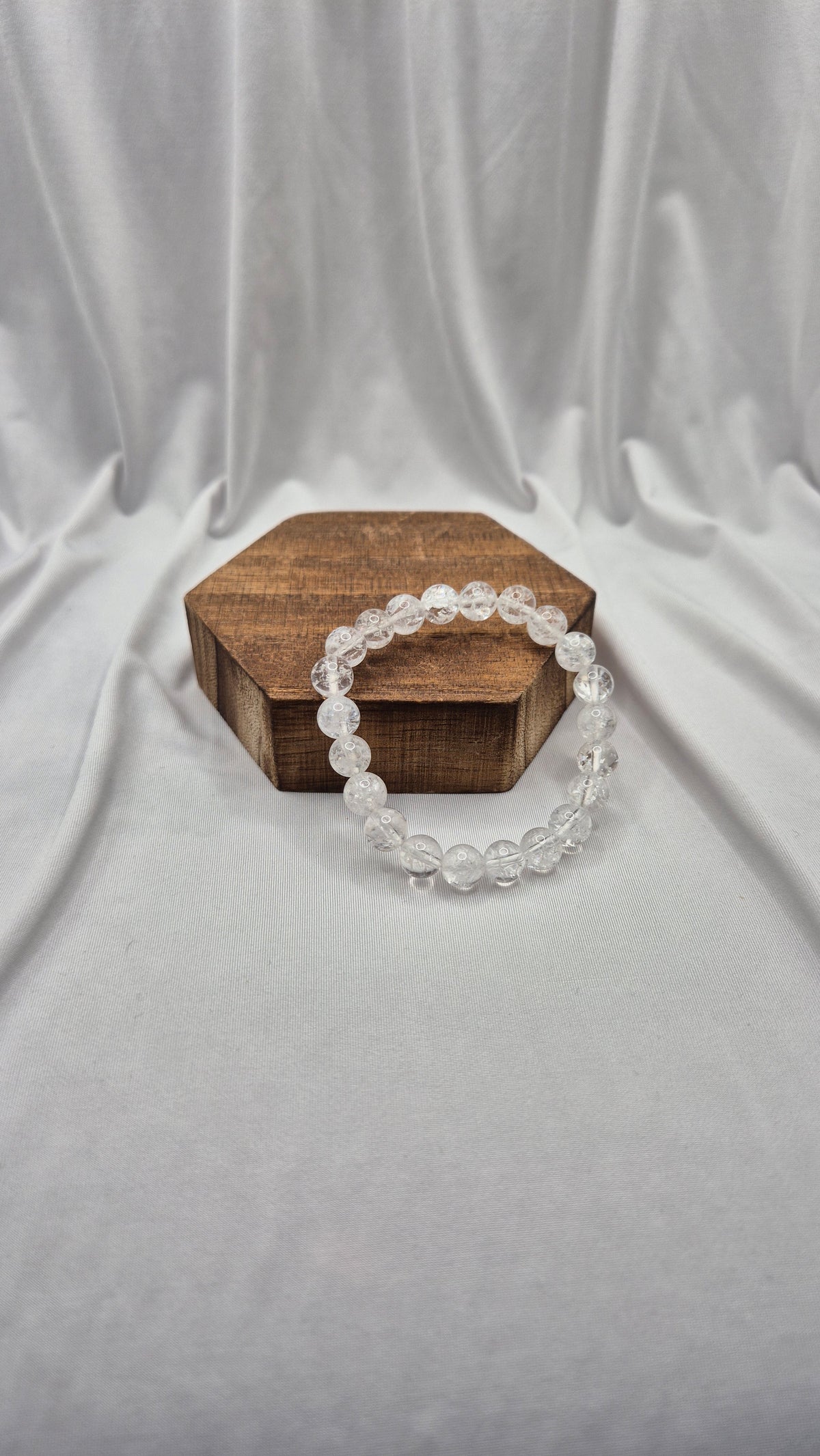 A clear beaded bracelet made of round transparent beads, draped over a small hexagonal wooden box on a draped white fabric backdrop.