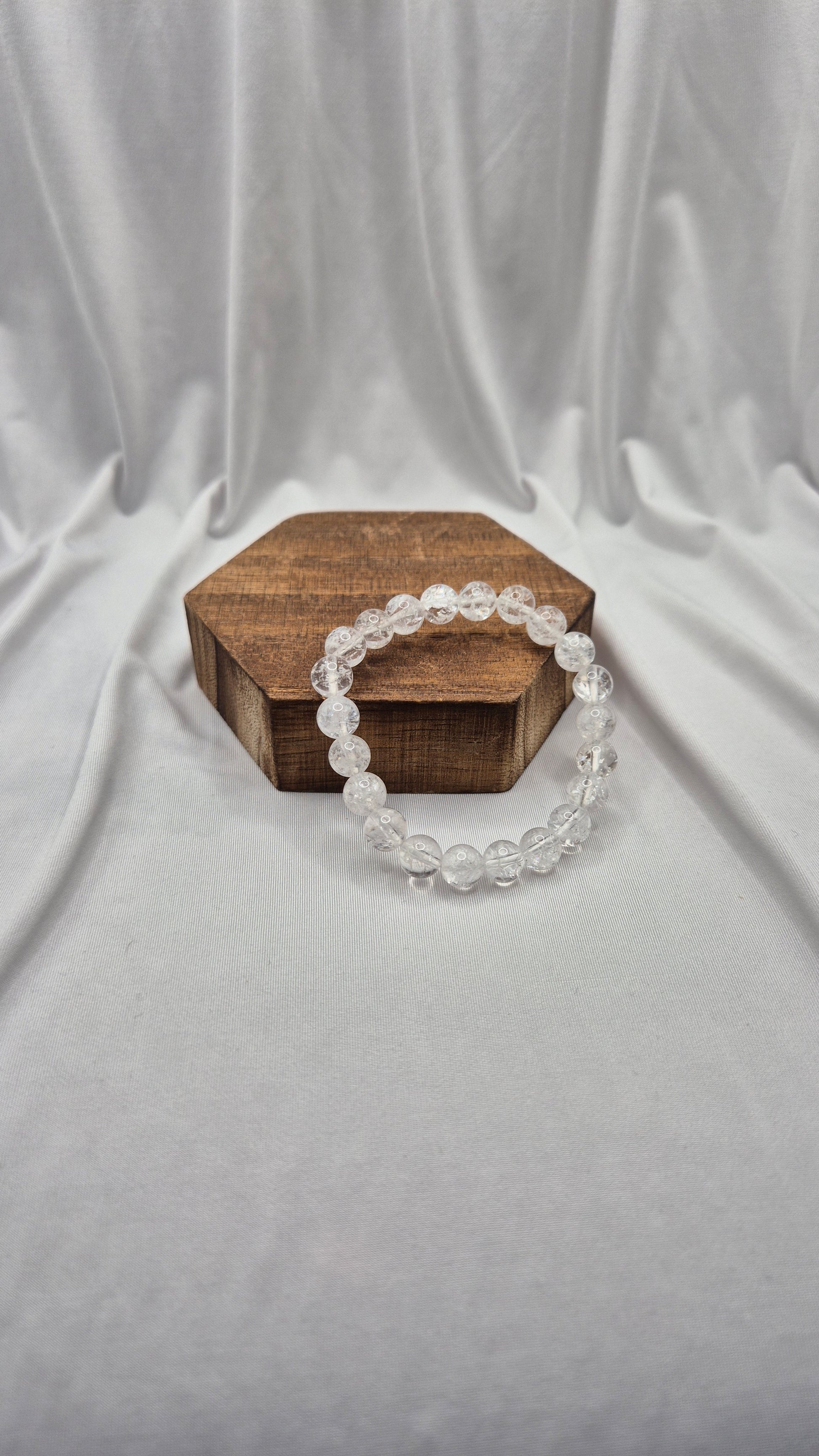 A clear beaded bracelet made of round transparent beads, draped over a small hexagonal wooden box on a draped white fabric backdrop.