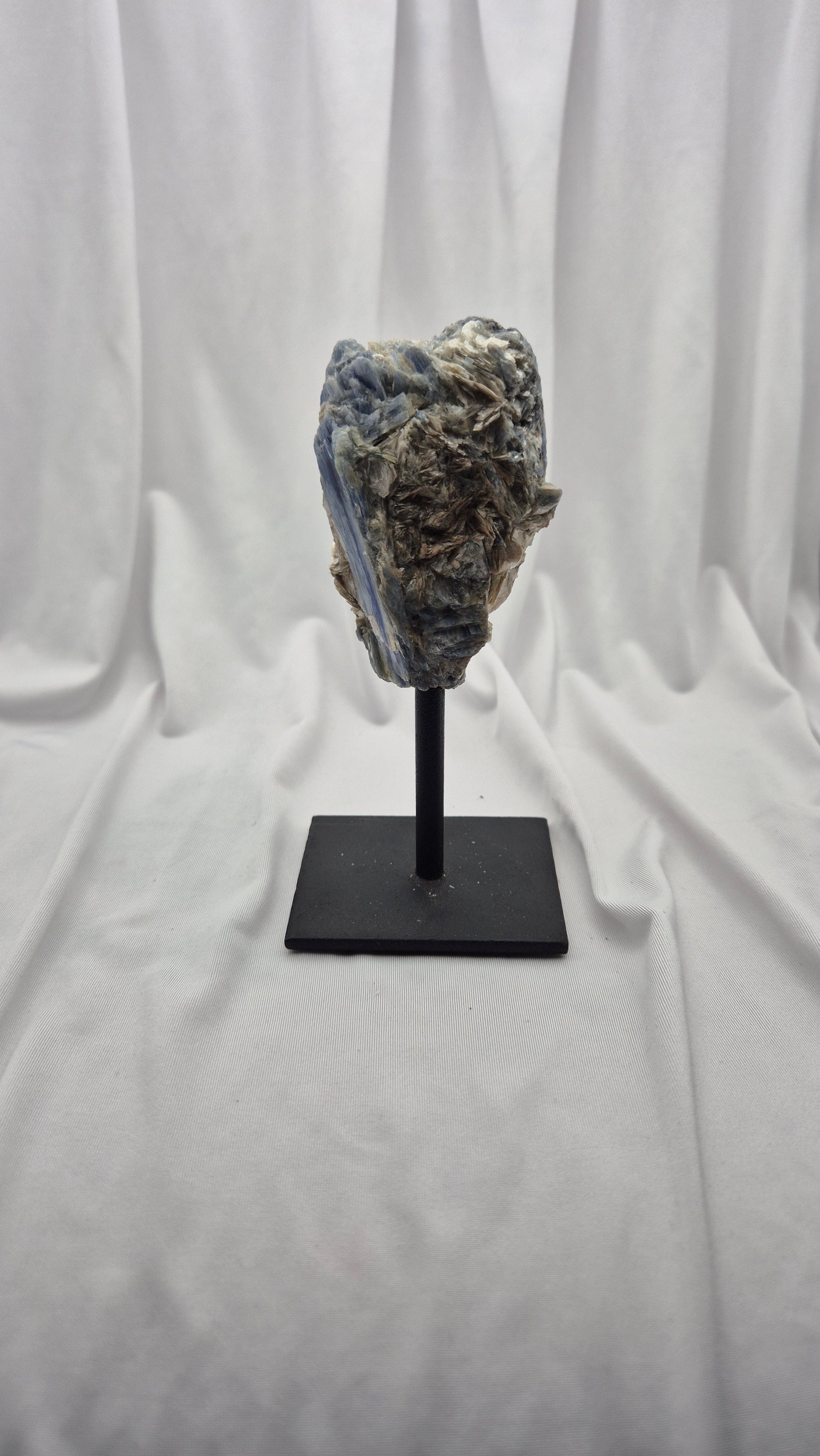 A rough blue agate crystal cluster mounted on a black display stand, set against a draped white fabric backdrop.