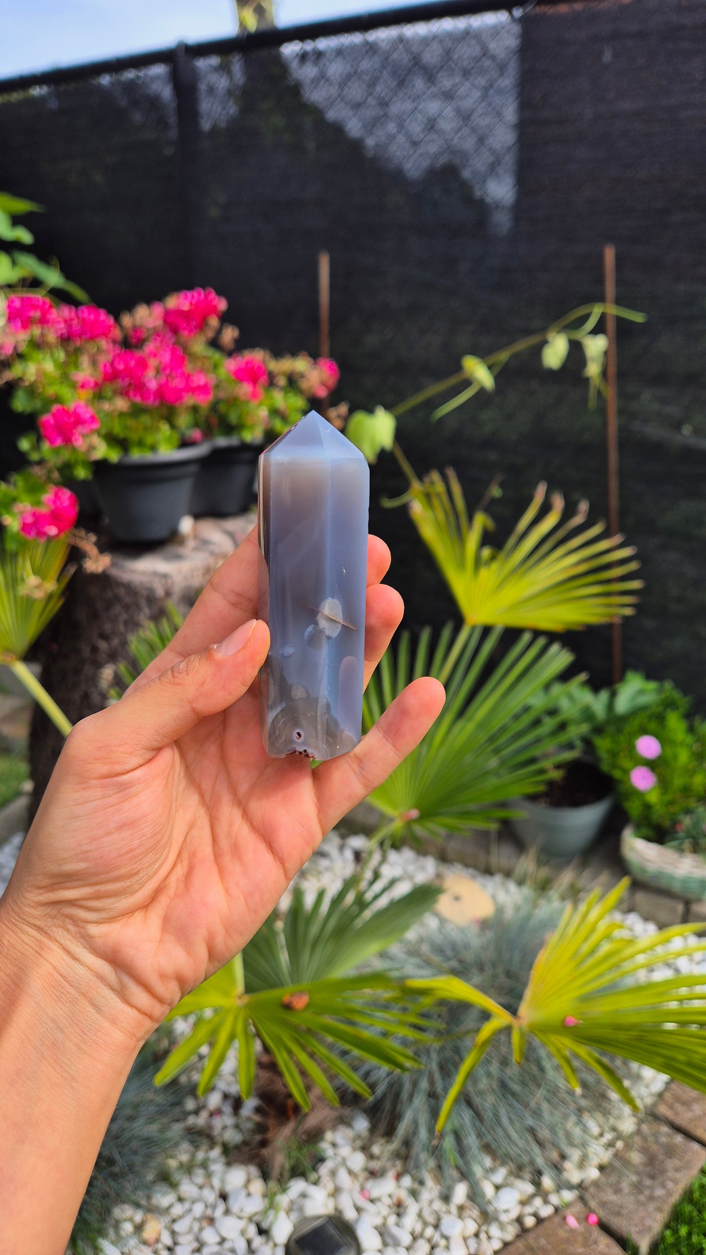 A hand holds a polished gray agate crystal point tower outdoors in a garden setting with potted pink flowers and green plants in the background.