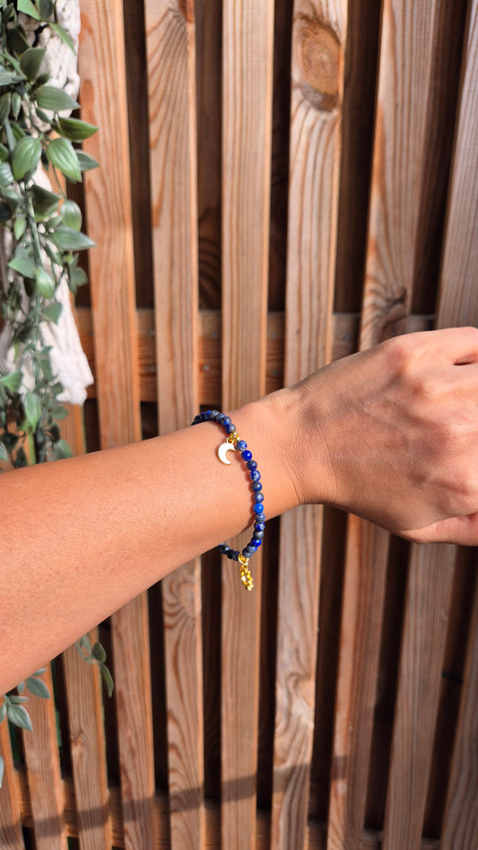 A close-up of a person's wrist wearing a beaded bracelet with dark blue beads, a small gold crescent moon charm, and a gold star accent, set against a wooden slat background.