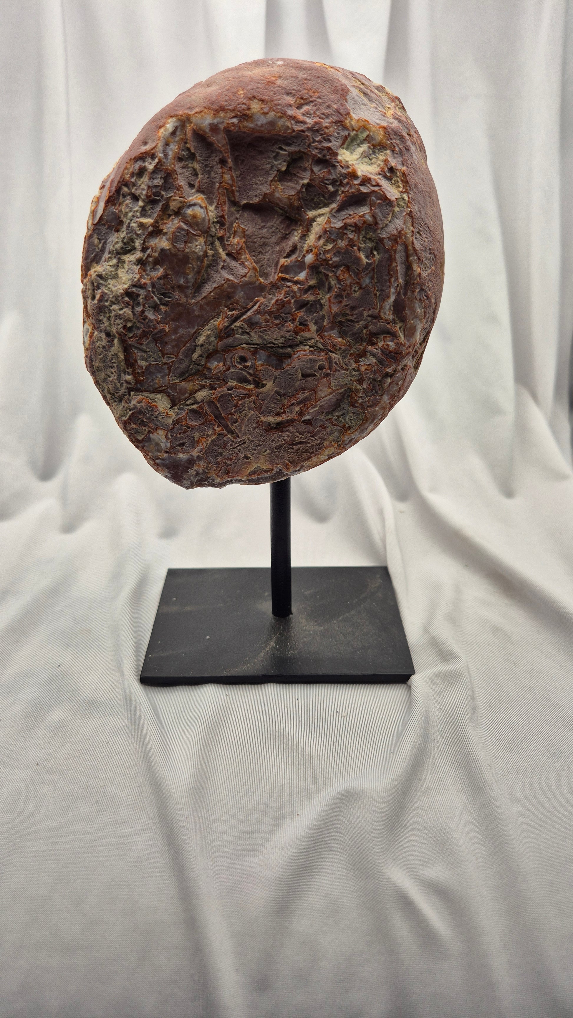 A textured brown stone or rock specimen mounted on a black display stand against a draped white fabric backdrop.