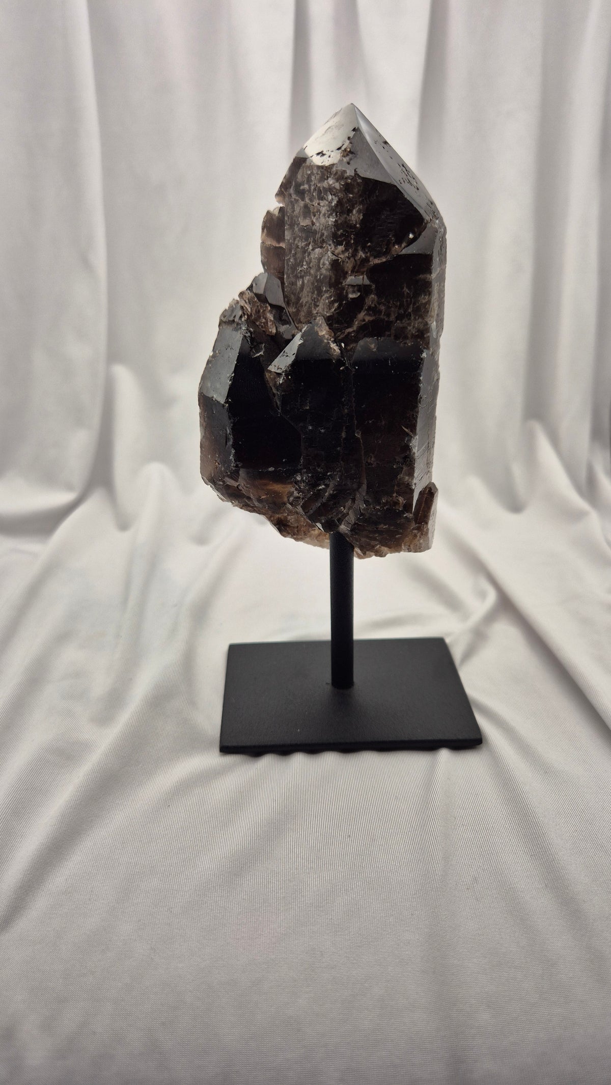 A dark brown to black crystal cluster on a black metal stand, photographed against a draped white fabric backdrop.