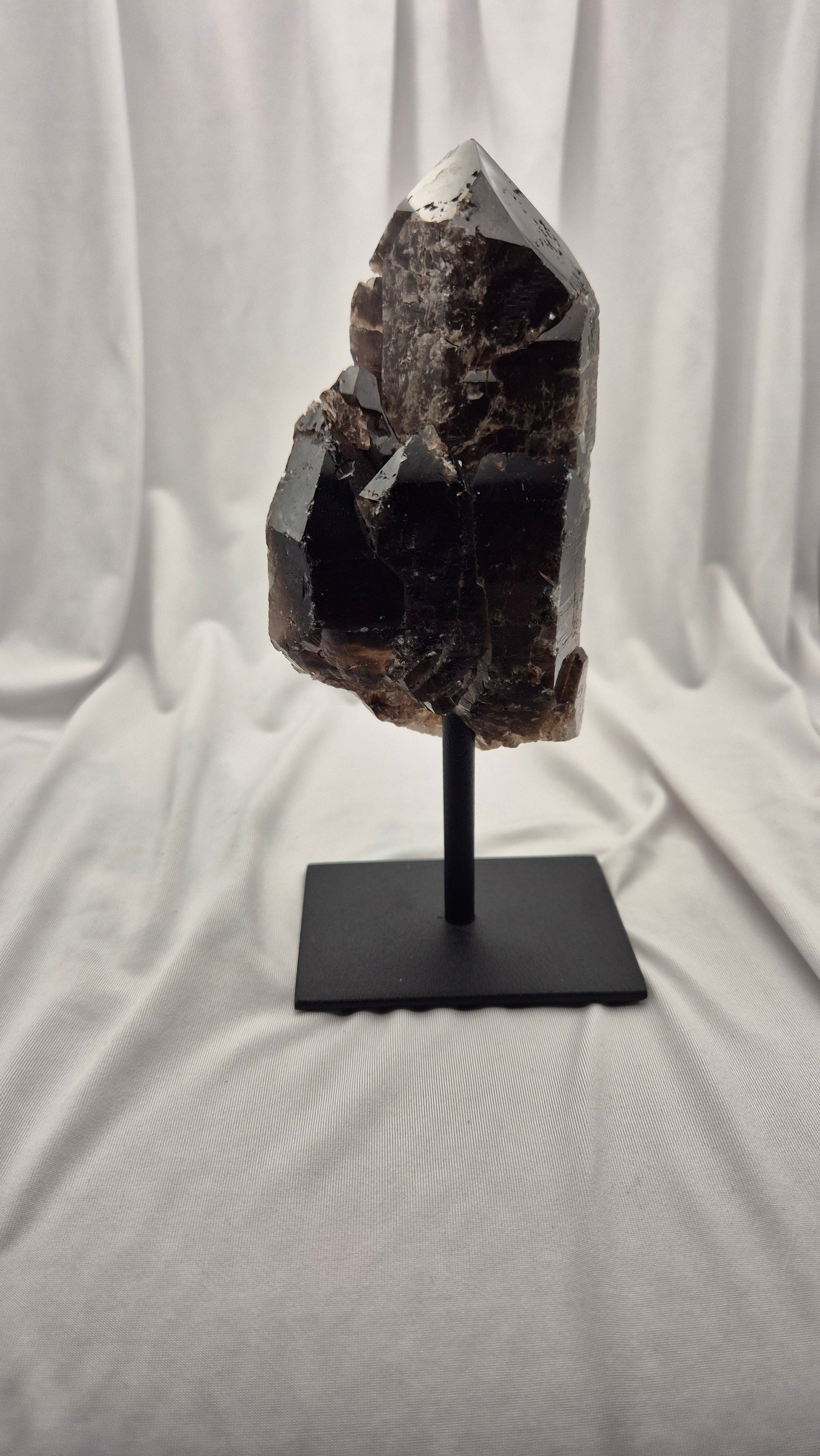 A dark brown to black crystal cluster on a black metal stand, photographed against a draped white fabric backdrop.