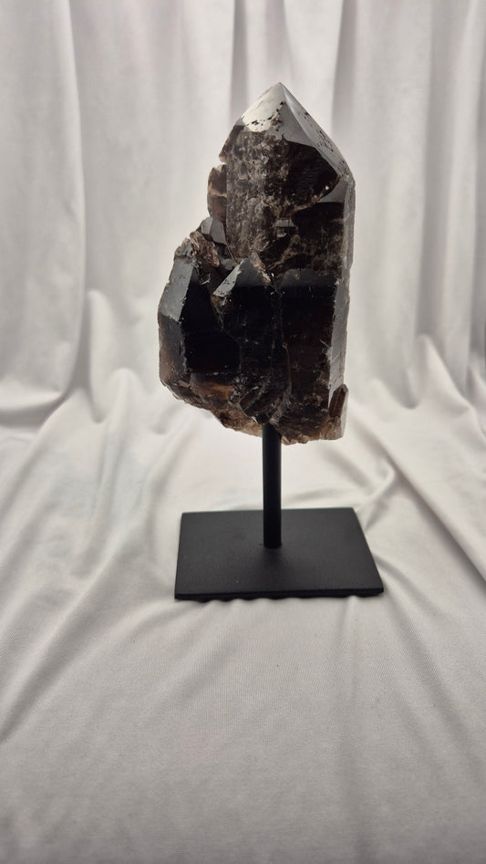 A dark brown to black crystal cluster on a black metal stand, photographed against a draped white fabric backdrop.