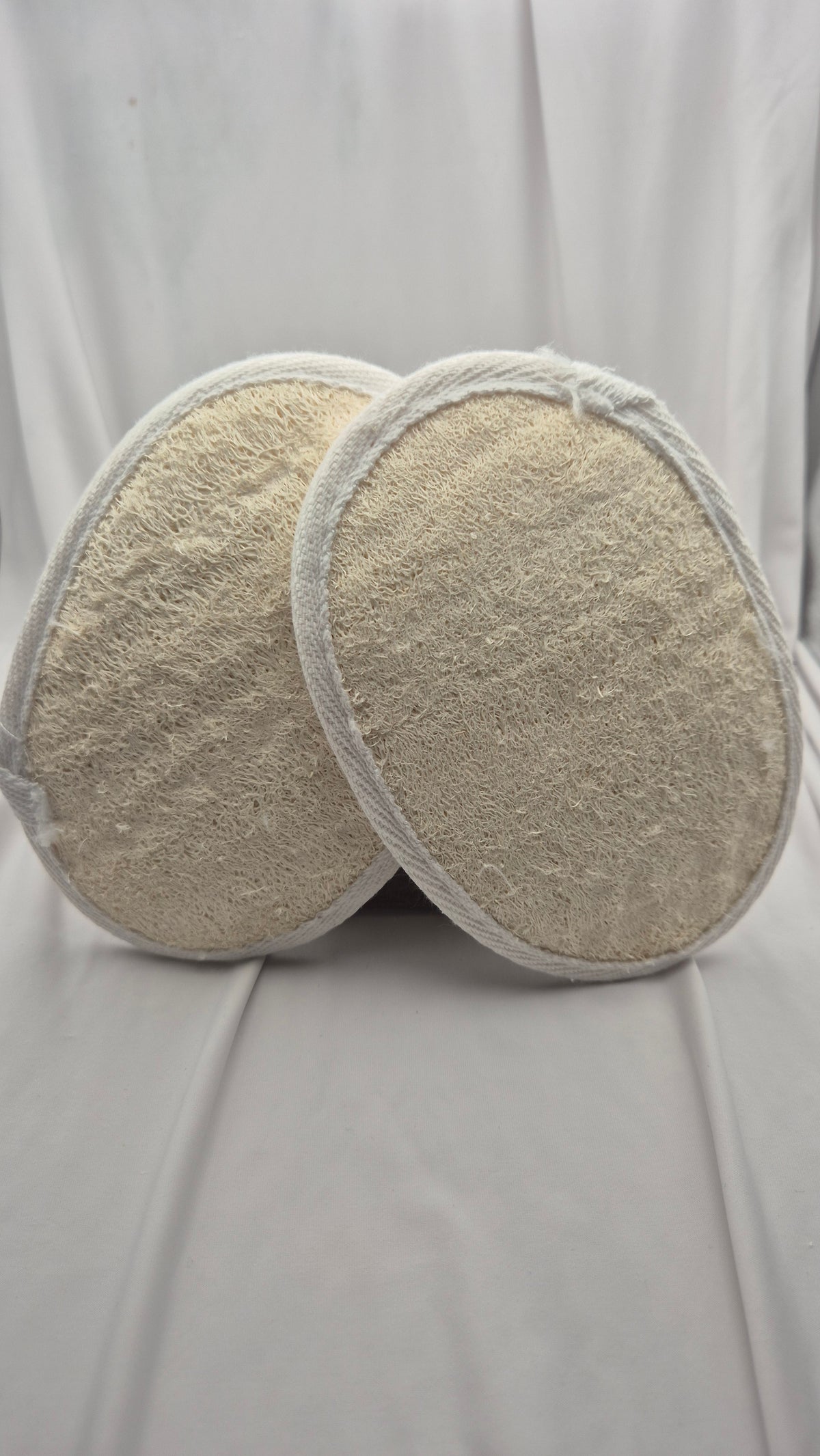 Two oval-shaped beige textured bath towels with white stitched edges, displayed overlapping on a draped white fabric background.