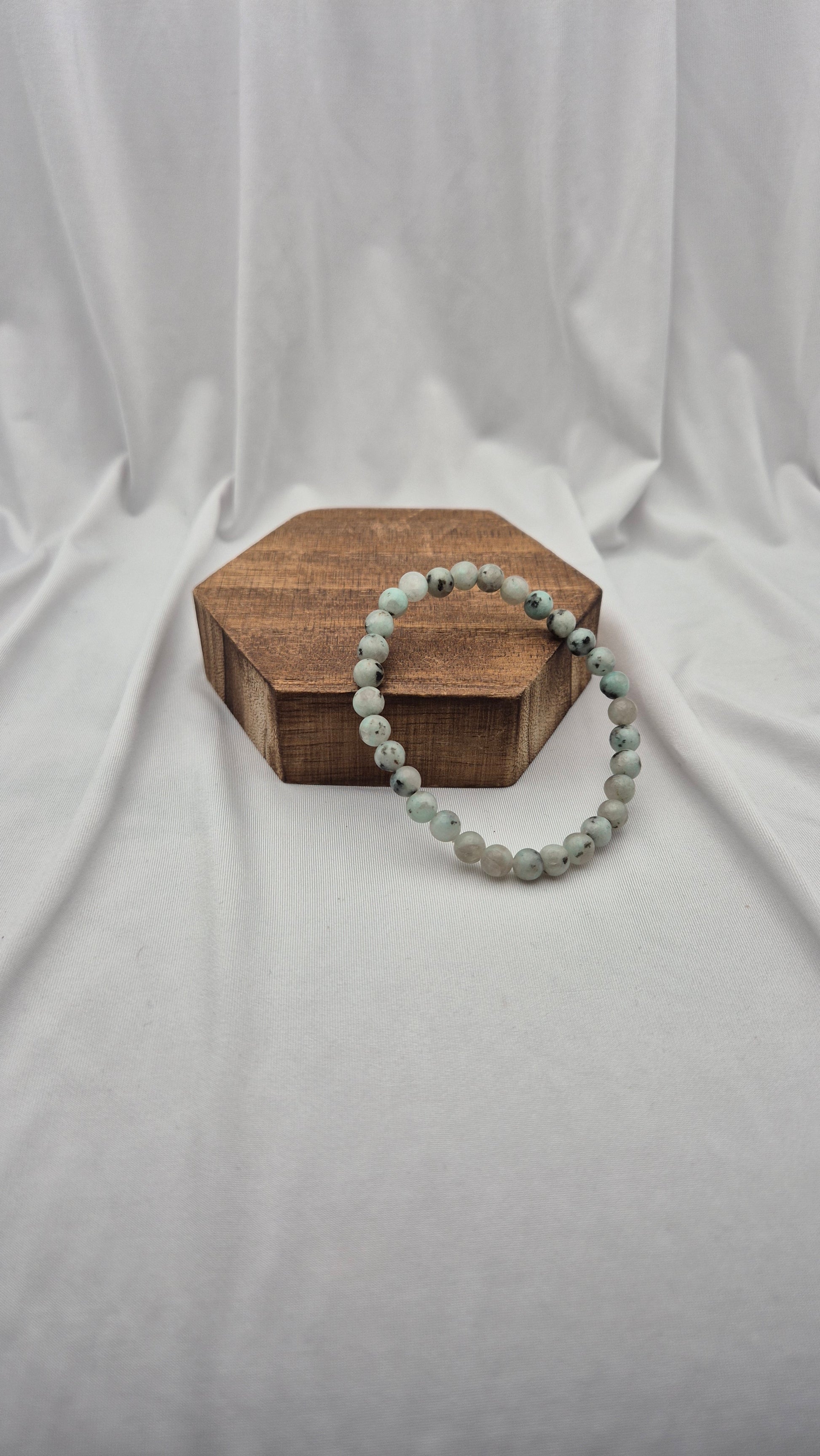 A beaded stretch bracelet made of light gray-green marbled beads, draped over a hexagonal wooden block on a draped white fabric background.