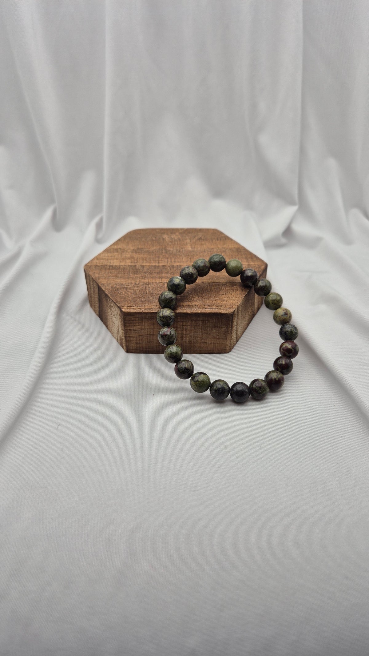 A beaded stretch bracelet made of dark, polished round beads with natural green and black marbling, displayed on a small hexagonal wooden block against a draped white fabric backdrop.