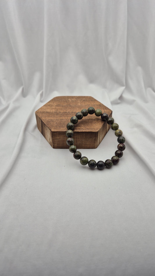 A beaded stretch bracelet made of dark, polished round beads with natural green and black marbling, displayed on a small hexagonal wooden block against a draped white fabric backdrop.