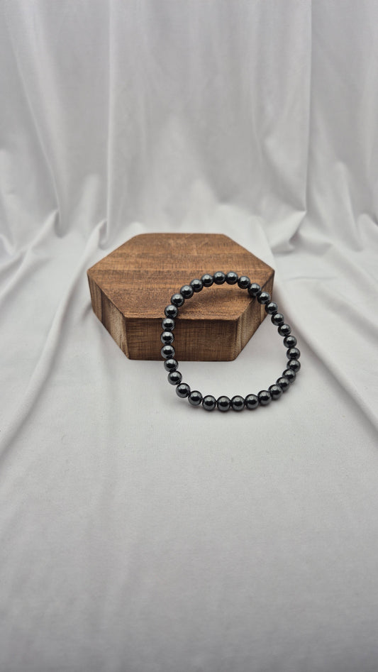 A black beaded bracelet displayed on a small hexagonal wooden block against a draped white fabric backdrop.