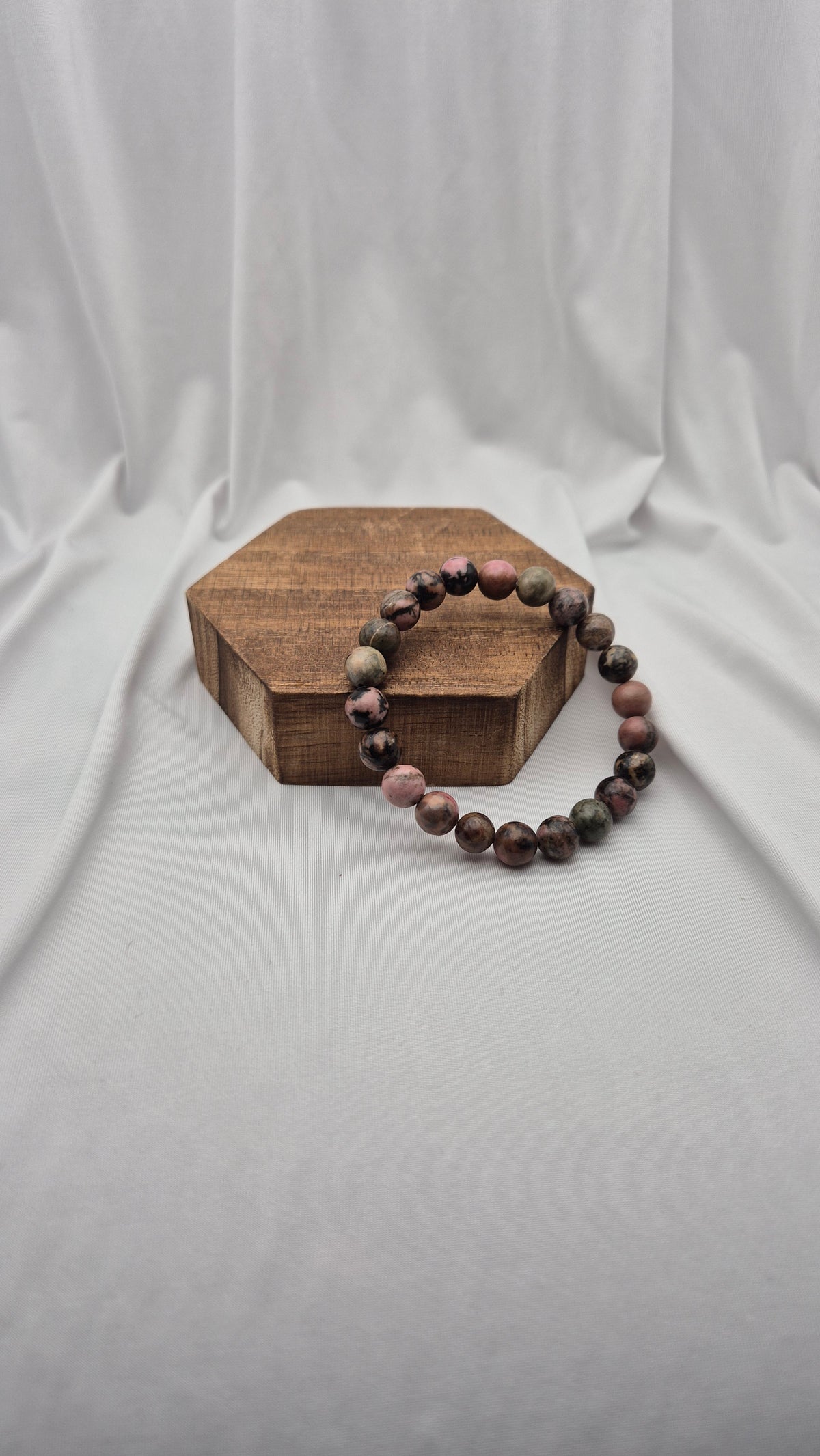 A beaded stretch bracelet made of polished round beads in earthy brown, gray, and pink tones, displayed on a small hexagonal wooden block against a draped white fabric backdrop.