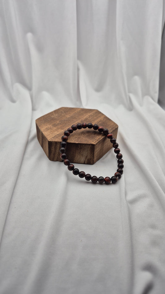 A dark brown beaded bracelet with small polished round beads, draped over a hexagonal wooden block on a draped white fabric background.