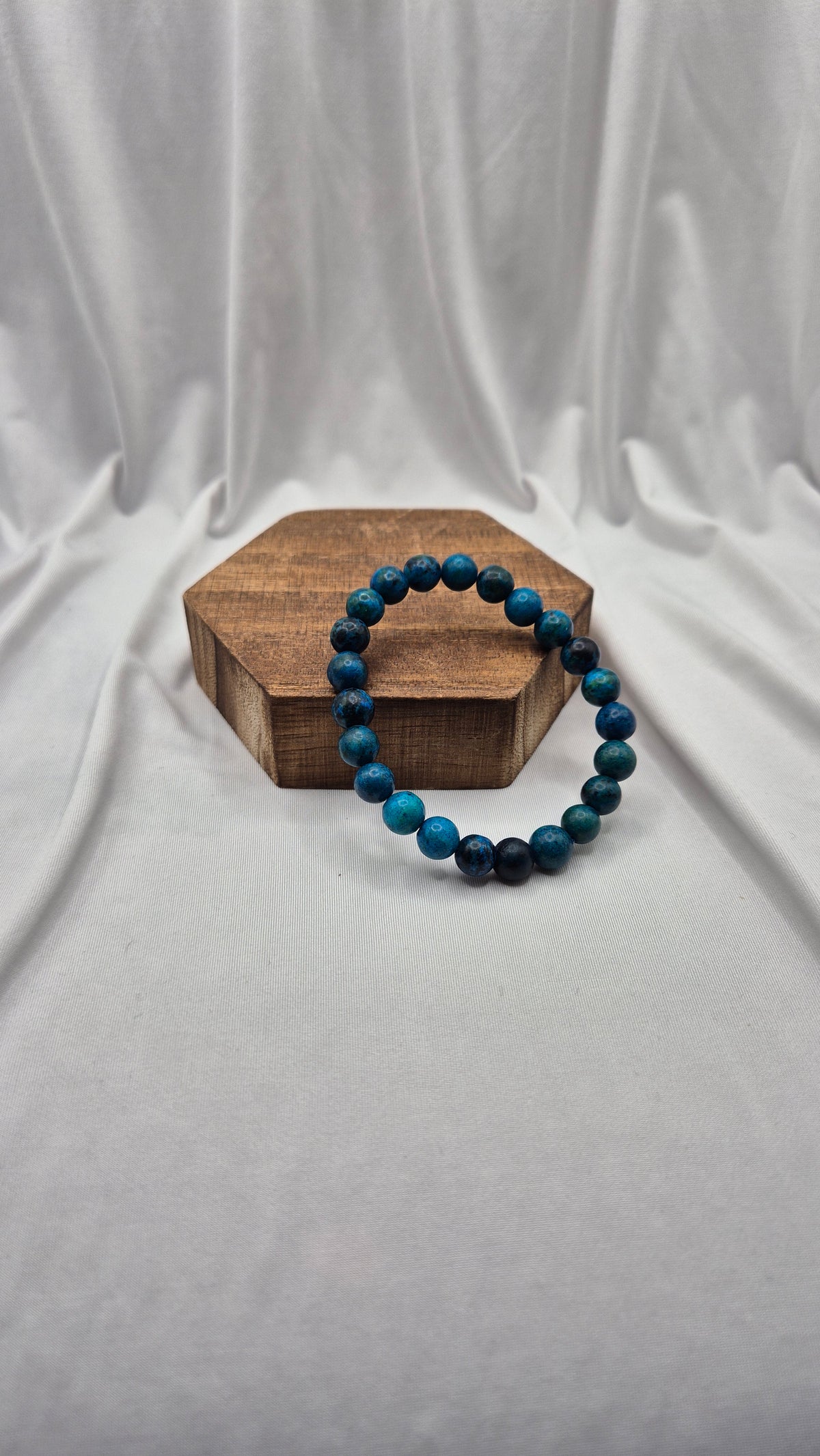 A beaded stretch bracelet made of dark blue and black beads, displayed on a small hexagonal wooden block against a draped white fabric backdrop.