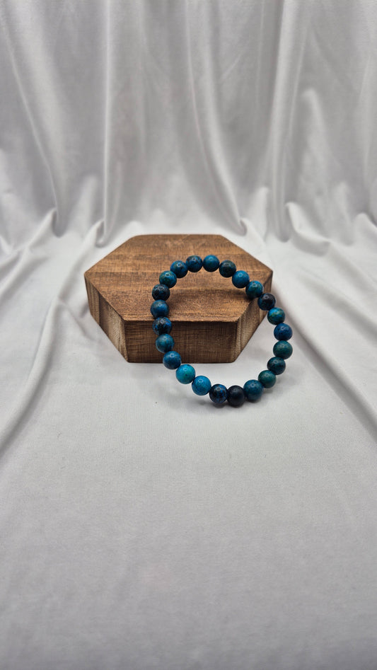 A beaded stretch bracelet made of dark blue and black beads, displayed on a small hexagonal wooden block against a draped white fabric backdrop.