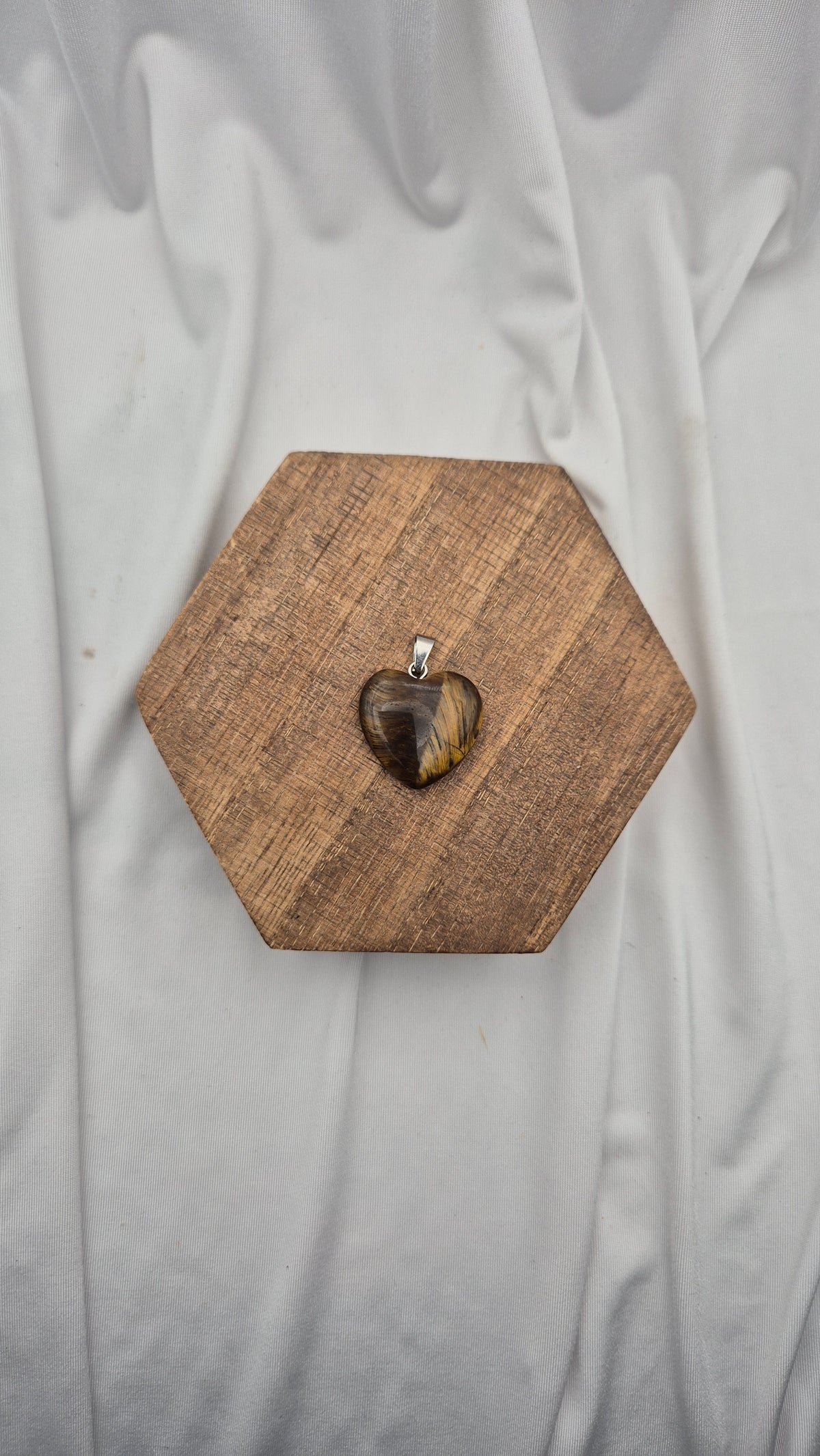 A heart-shaped pendant with a tiger's eye pattern, displayed on a hexagonal wooden block against a draped white fabric background.