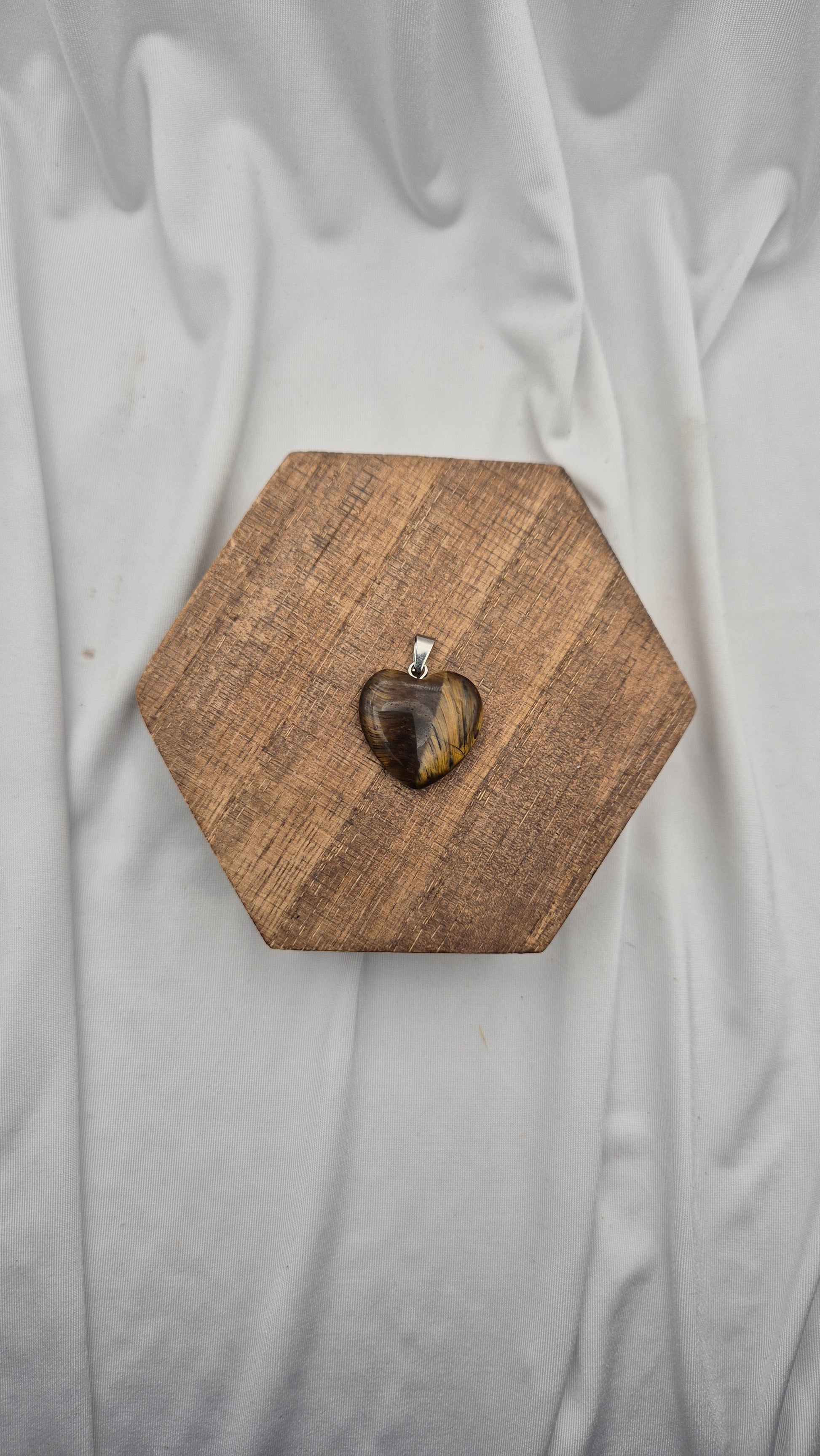 A heart-shaped pendant with a tiger's eye pattern, displayed on a hexagonal wooden block against a draped white fabric background.