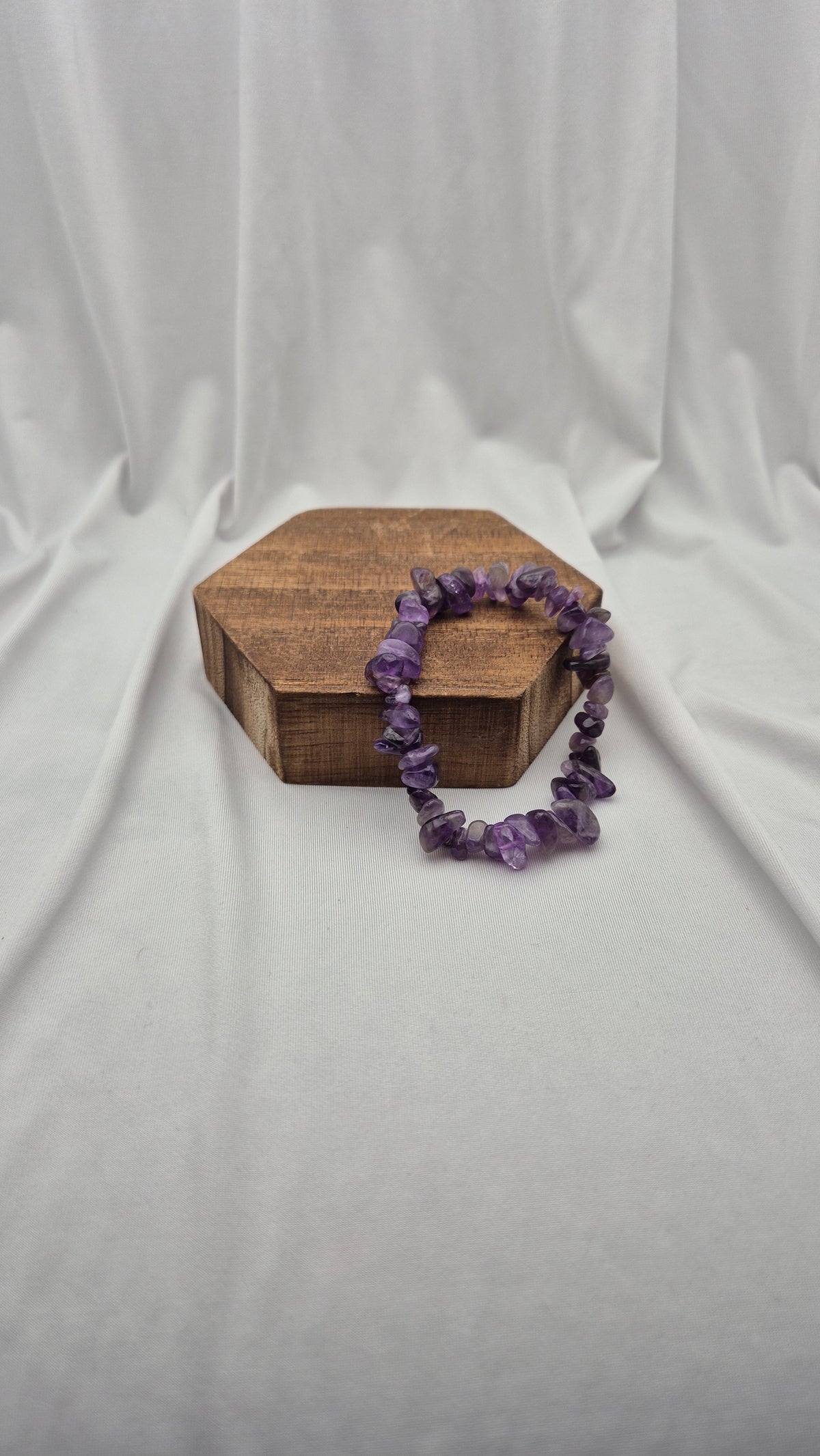 A purple amethyst chip bracelet draped over a small hexagonal wooden box, set against a draped white fabric backdrop.