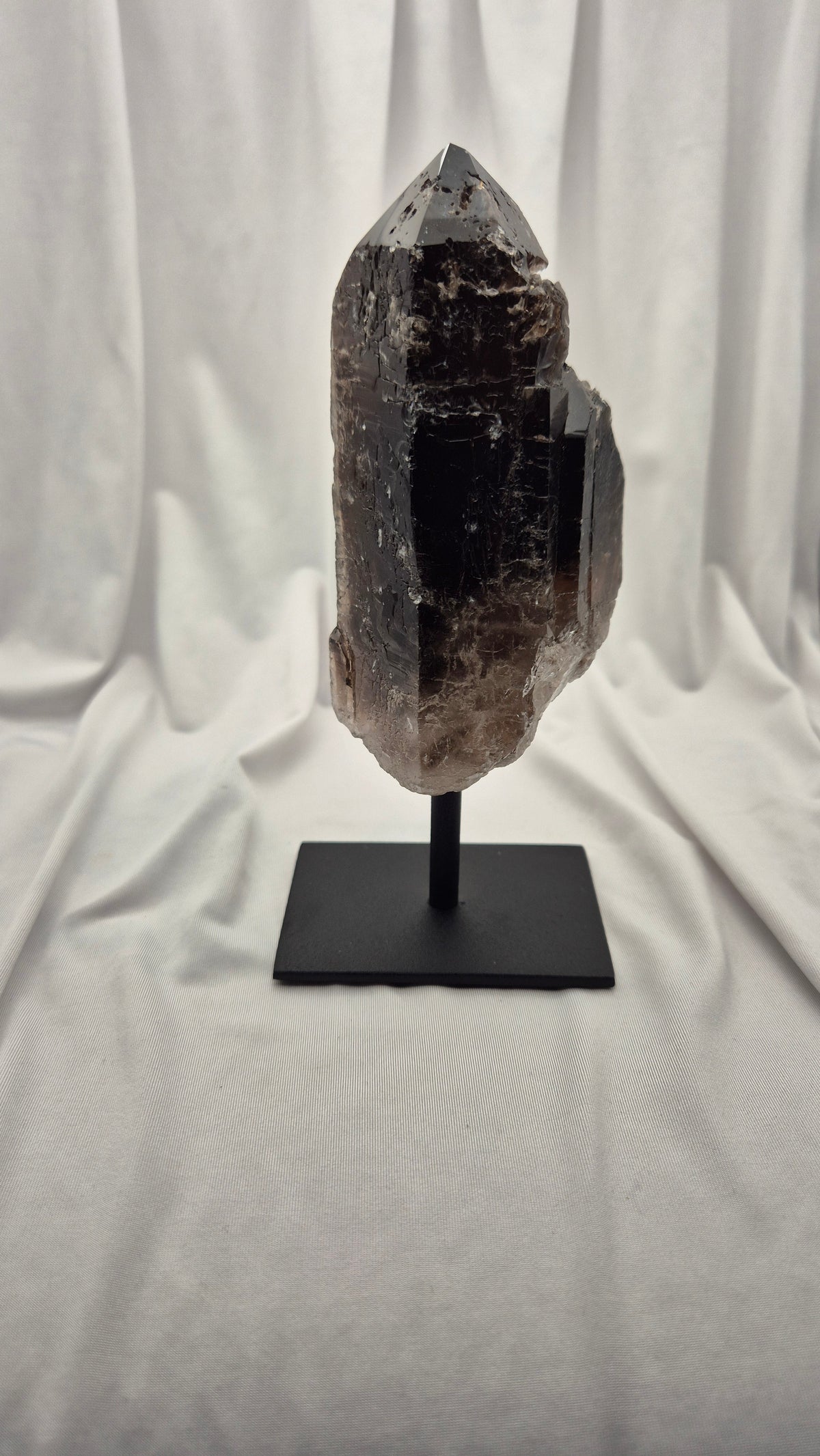 A raw smoky quartz crystal mount on a black metal display stand, set against a draped white fabric backdrop.