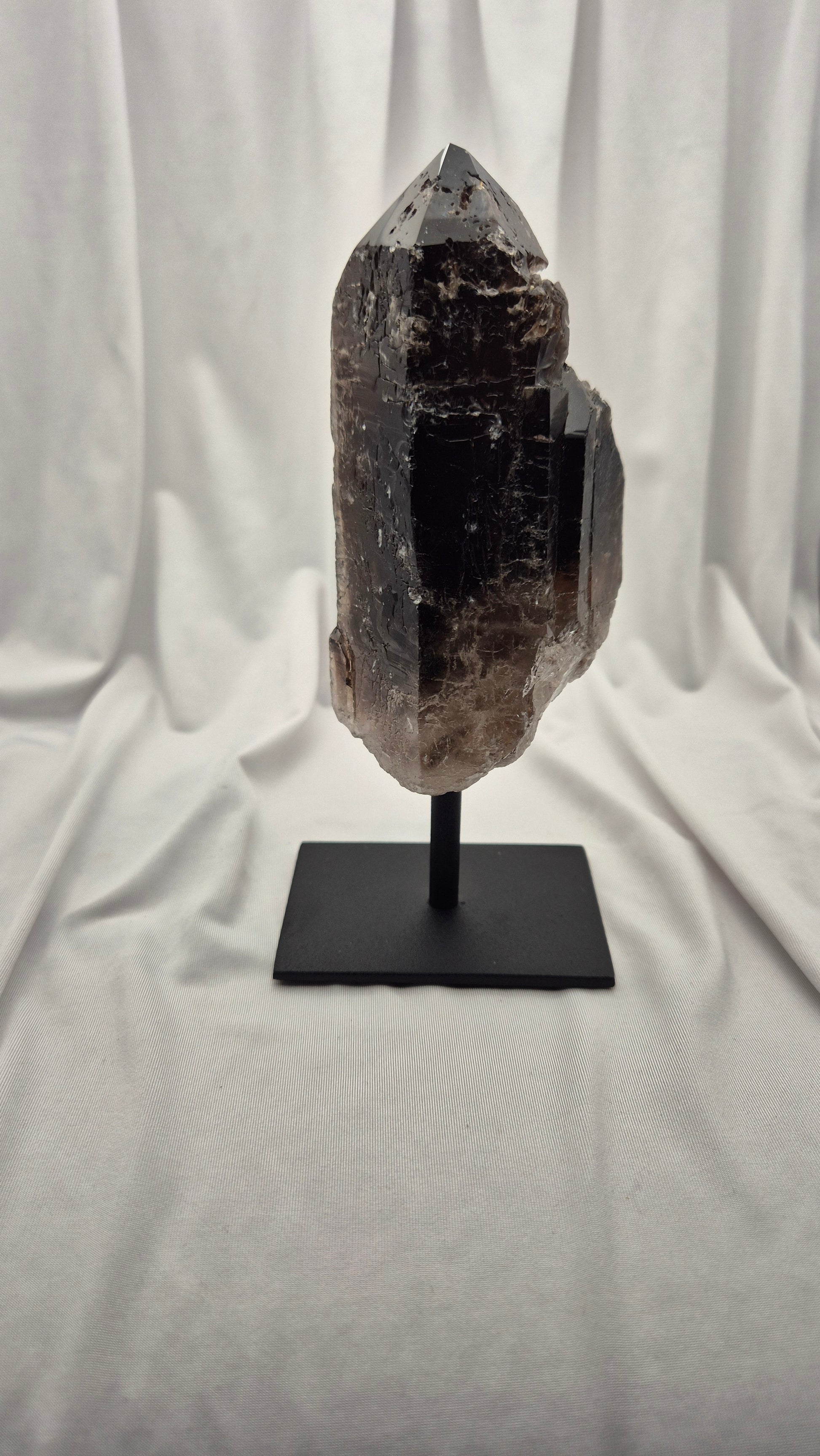 A raw smoky quartz crystal mount on a black metal display stand, set against a draped white fabric backdrop.