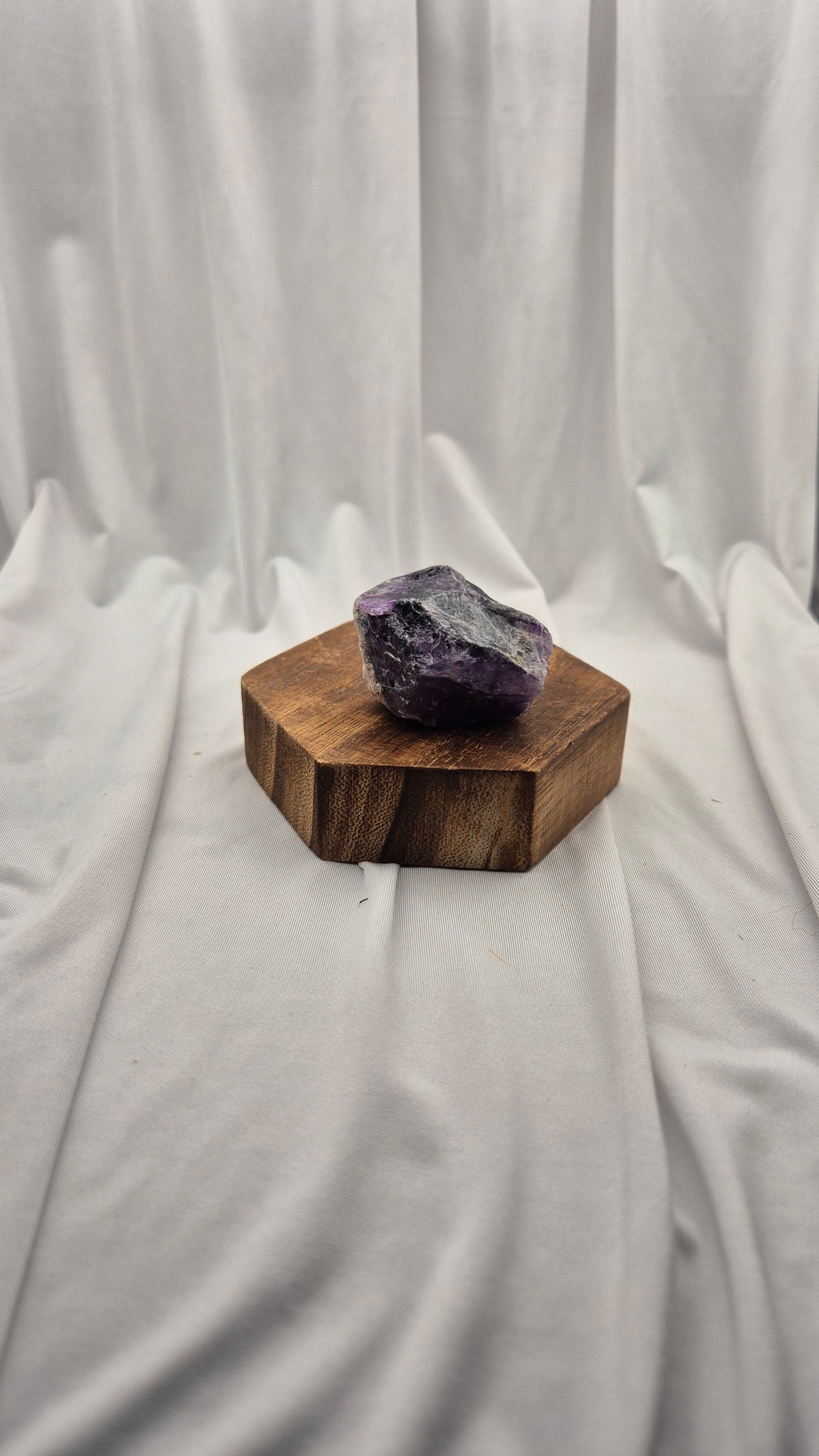 A raw purple amethyst crystal resting on a hexagonal wooden block, set against a draped white fabric backdrop.