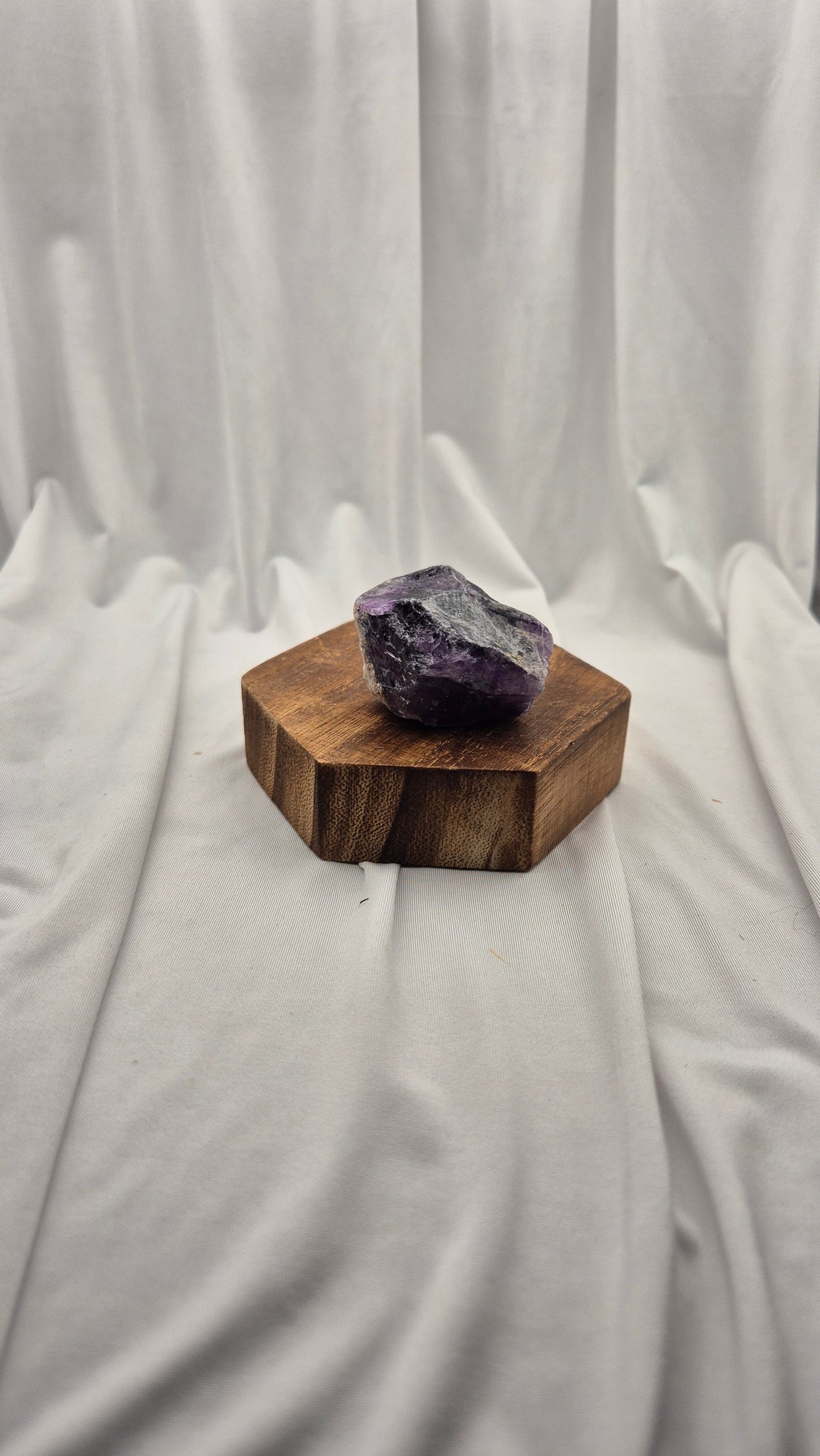 A raw purple amethyst crystal resting on a hexagonal wooden block, set against a draped white fabric backdrop.