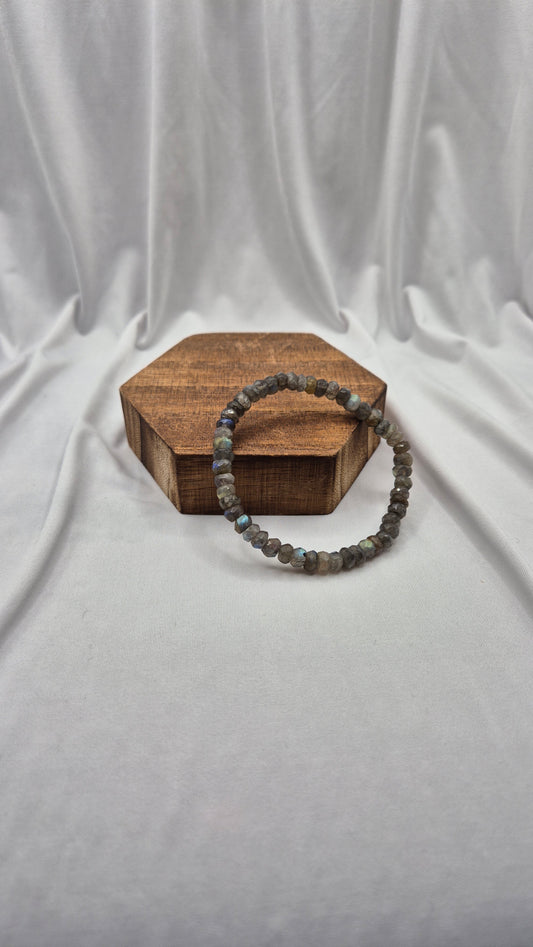 A beaded bracelet made of small gray and blue-green stones, draped over a hexagonal wooden box against a draped white fabric backdrop.