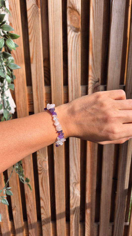 A person's wrist wearing a beaded bracelet made of purple and white stones, posed in front of a wooden slat fence.