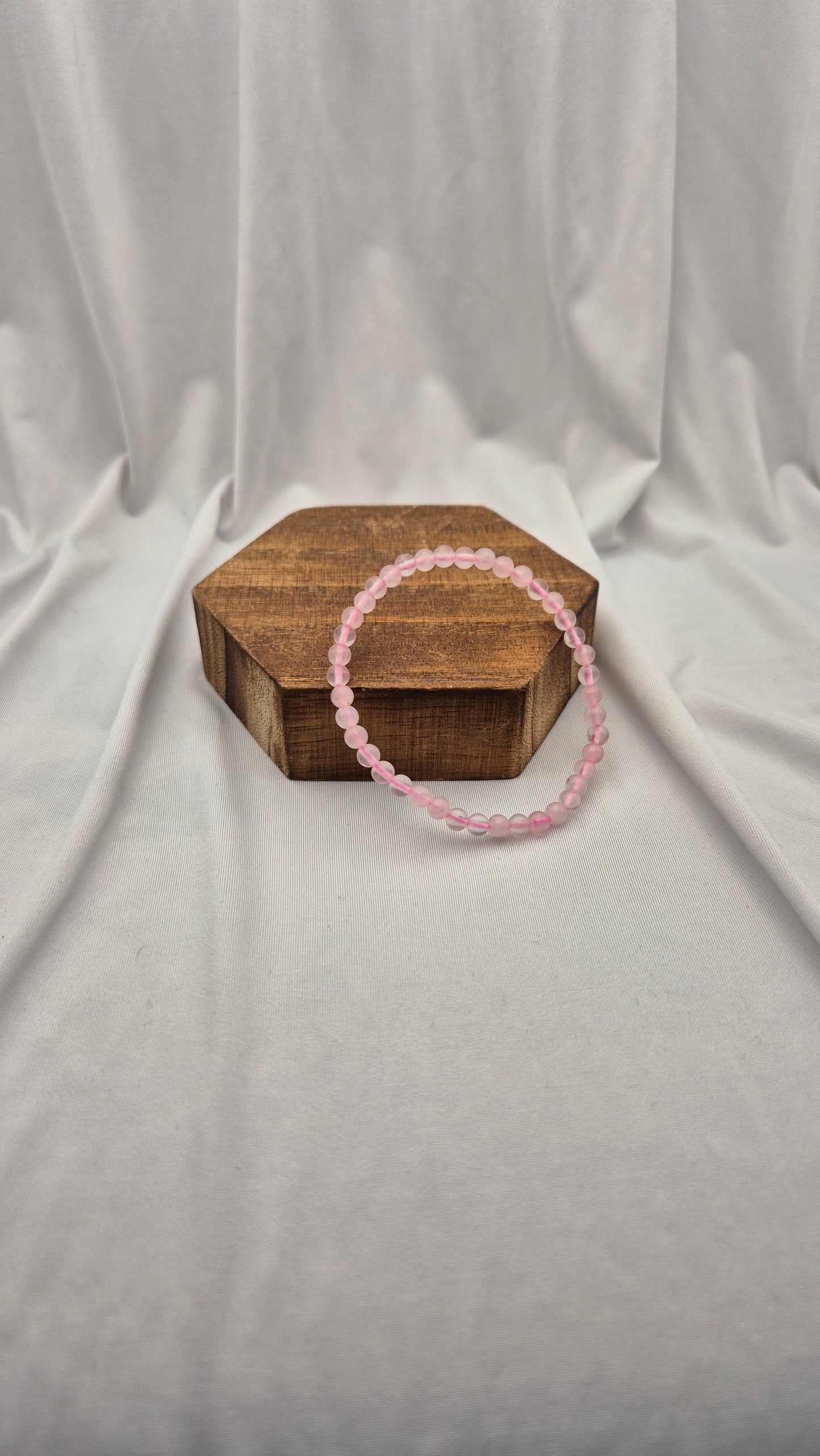 A pink beaded bracelet draped over a small hexagonal wooden box, set against a draped white fabric backdrop.