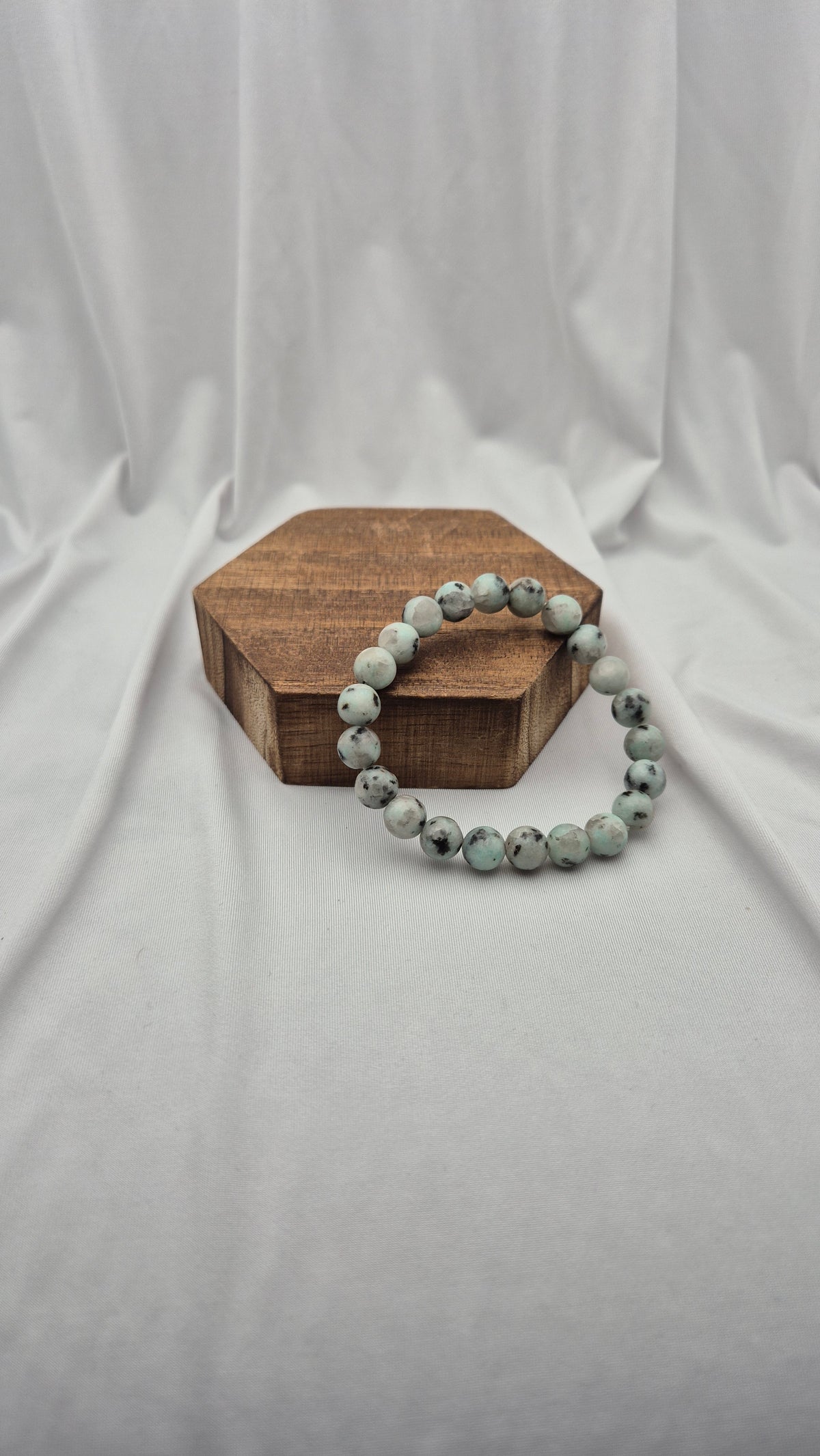 A beaded stretch bracelet made of round, speckled light gray-green beads resting on a small hexagonal wooden block against a draped white fabric backdrop.
