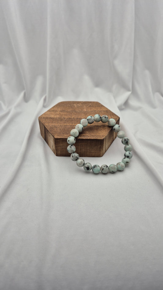 A beaded stretch bracelet made of round, speckled light gray-green beads resting on a small hexagonal wooden block against a draped white fabric backdrop.