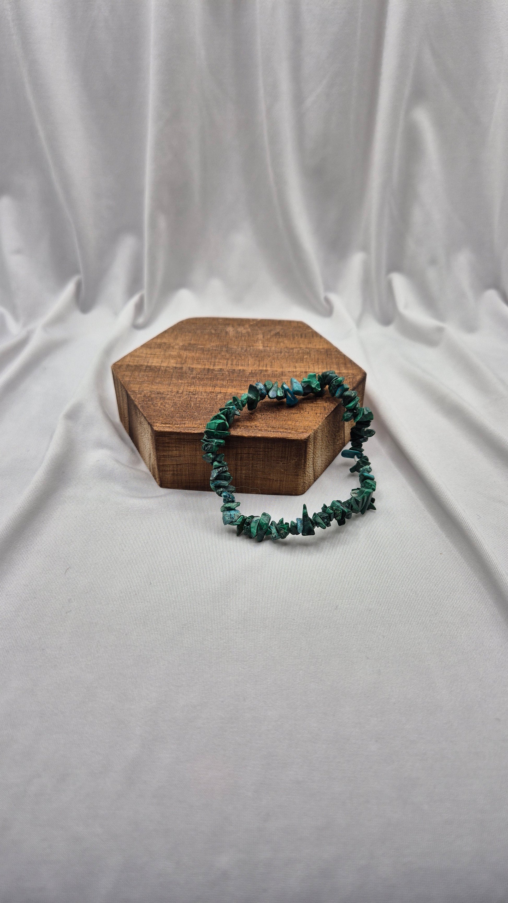 A green chunky stone bead bracelet draped over a small hexagonal wooden box, set against a draped white fabric backdrop.