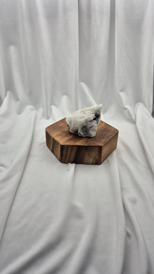 A raw white crystal cluster with dark inclusions sits on a hexagonal dark brown wooden block, set against a draped white fabric backdrop.