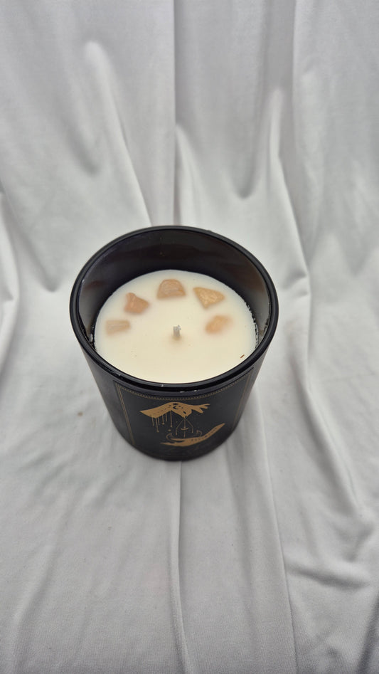 A black glass jar candle with a single wick, filled with white wax and decorated with small tan stone pieces on top, set against a draped white fabric background.