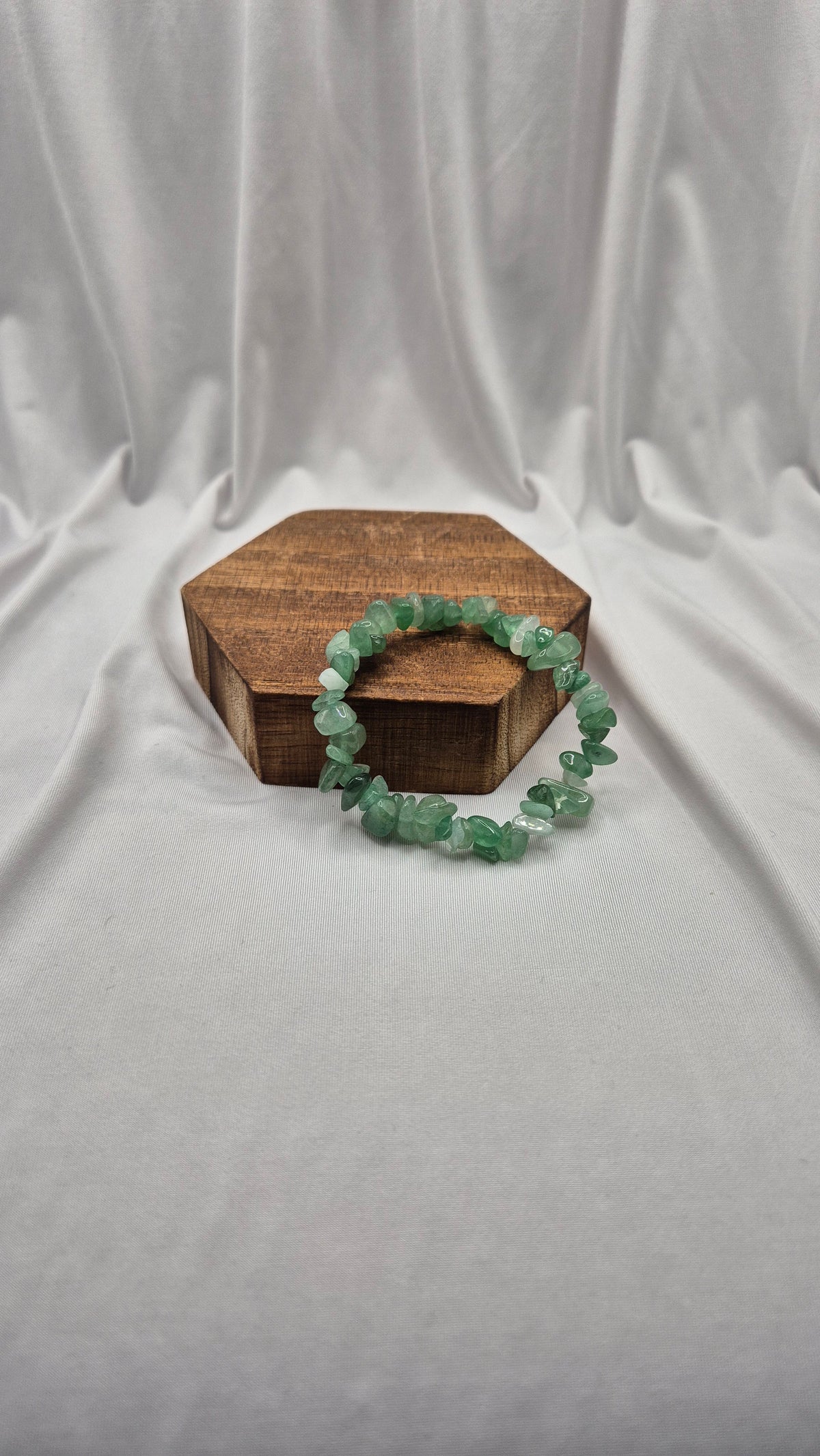 A green gemstone leaf beaded bracelet resting on a small hexagonal wooden box against a draped white fabric backdrop.