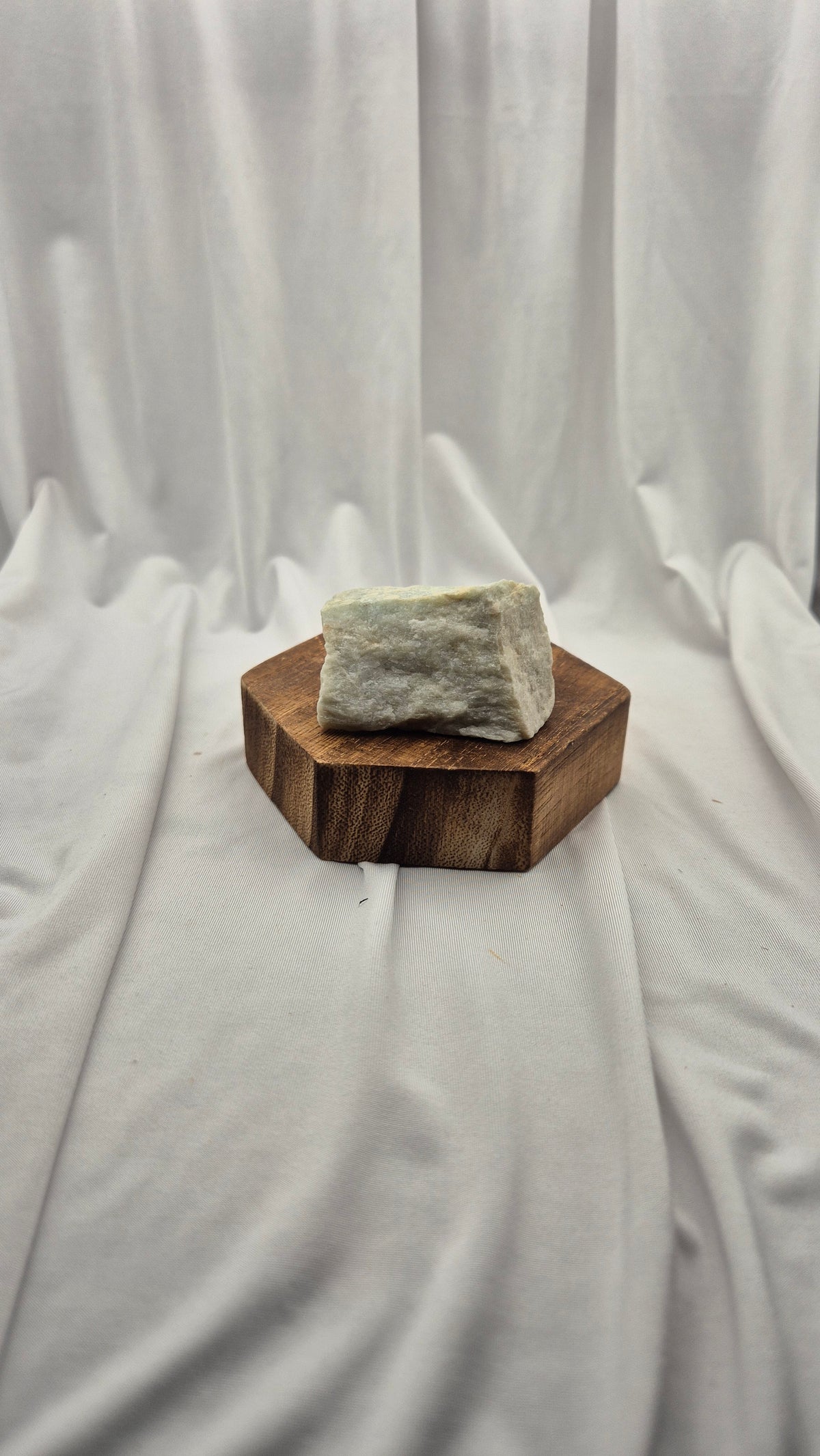 A rough, pale greenish-gray stone or crystal block displayed on a small hexagonal wooden stand against a draped white fabric backdrop.