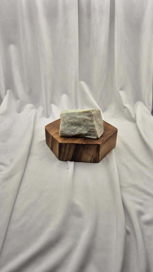 A rough, pale greenish-gray stone or crystal block displayed on a small hexagonal wooden stand against a draped white fabric backdrop.