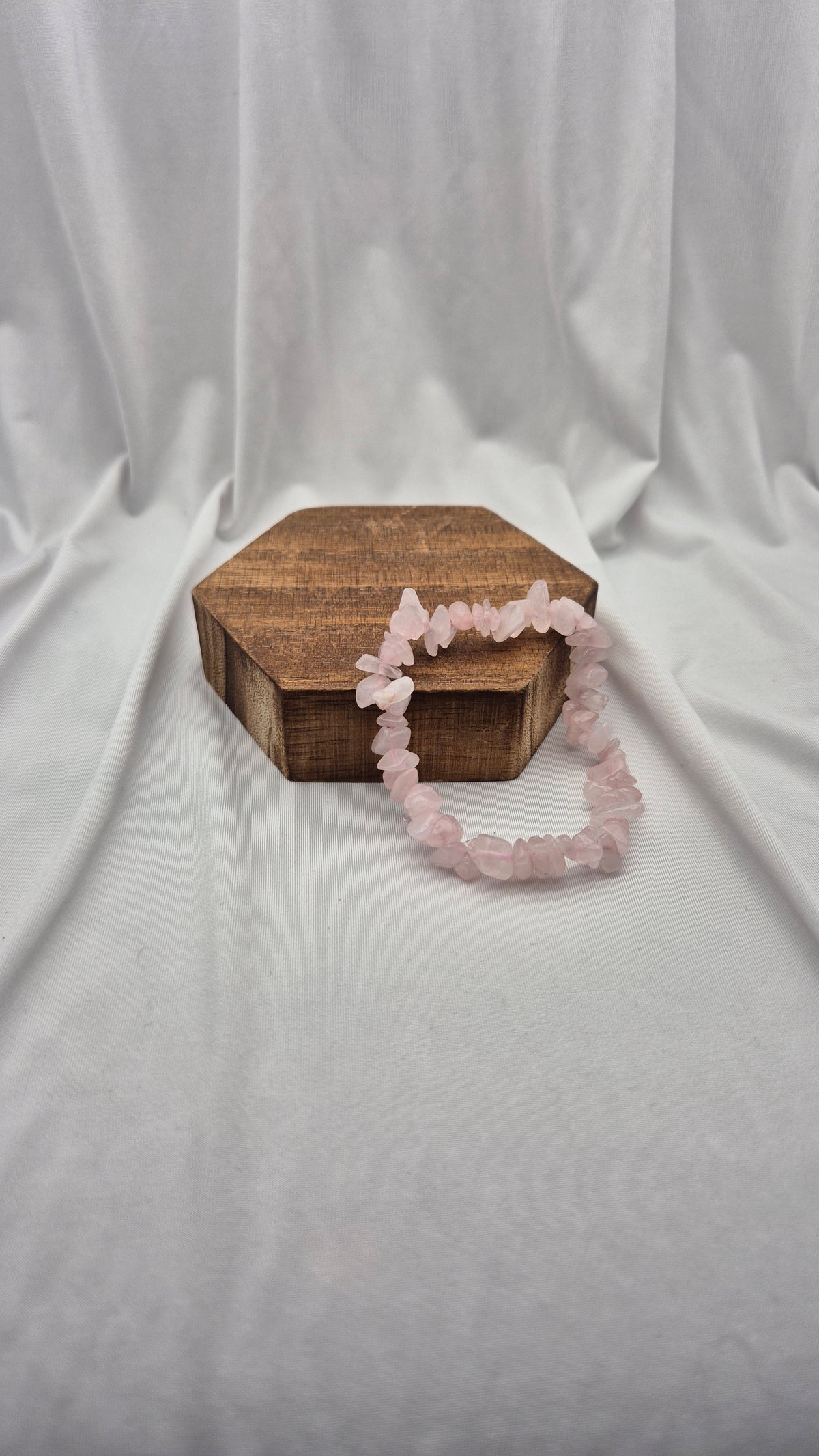 A pink rose quartz chip bracelet draped over a small hexagonal wooden box, set against a draped white fabric backdrop.