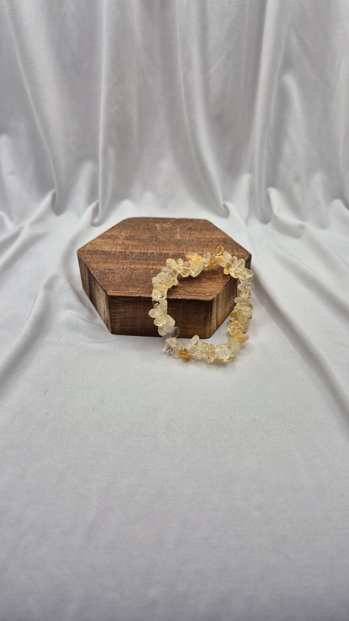 A beaded stretch bracelet made of irregular, translucent pale yellow and cream stones, draped over a small hexagonal wooden box on a draped white fabric background.